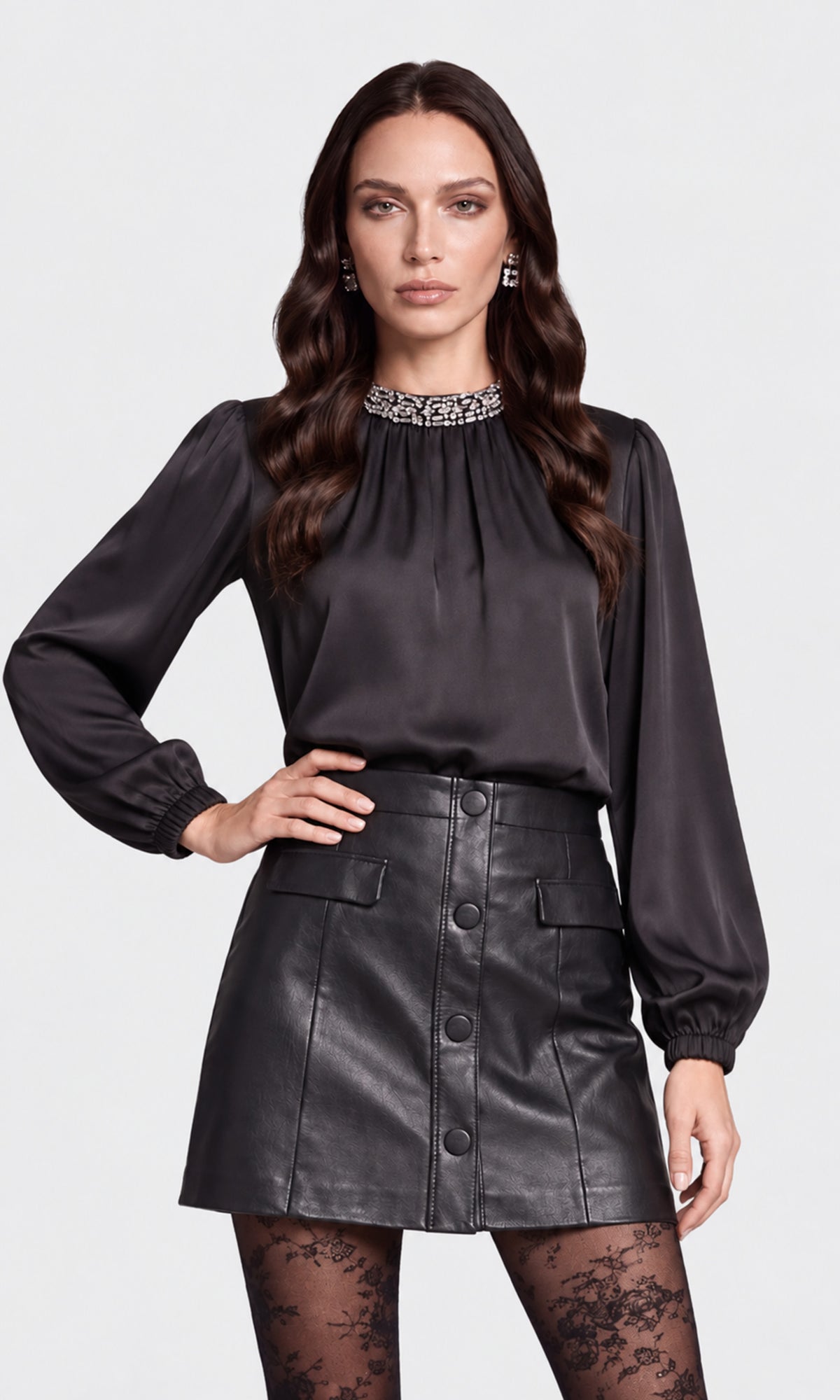 Model wearing a black long-sleeve satin blouse with a jeweled high neckline, paired with a black faux-leather button-front mini skirt and black lace tights.