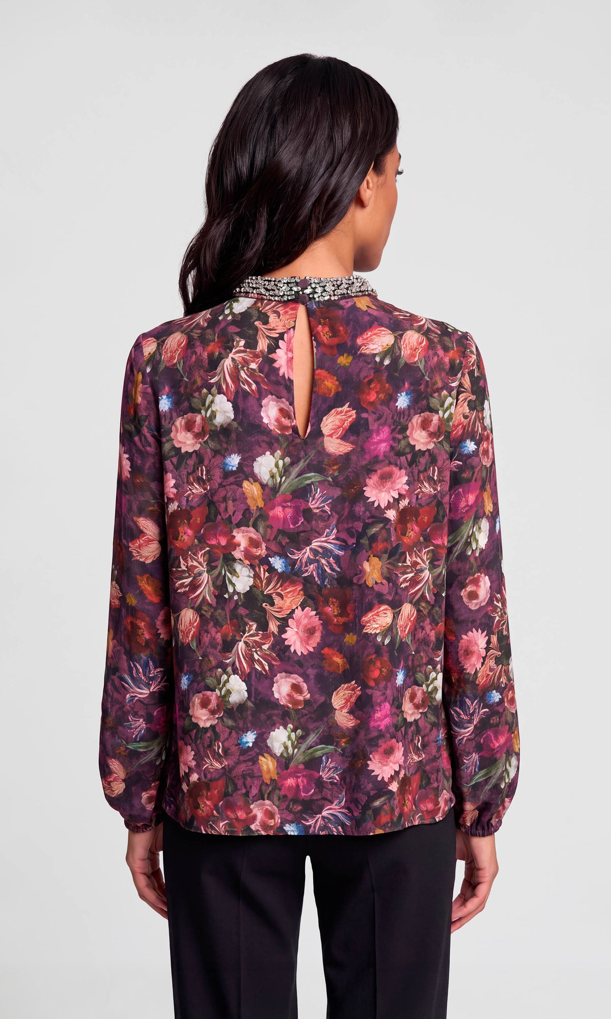 Back view of model wearing a long-sleeve floral blouse with a high gathered neckline and a beaded collar trim, paired with black pants.