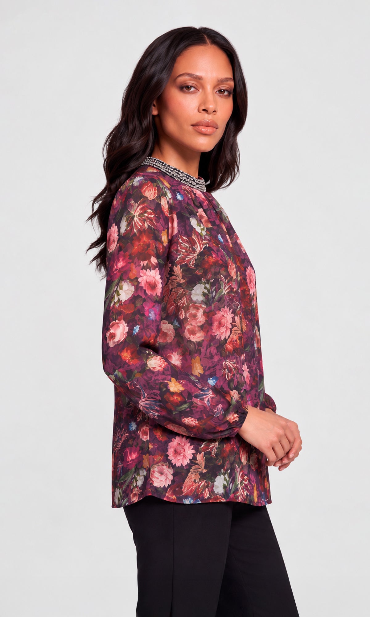 Side view of model wearing a long-sleeve floral blouse with a high gathered neckline and a beaded collar trim, paired with black pants.