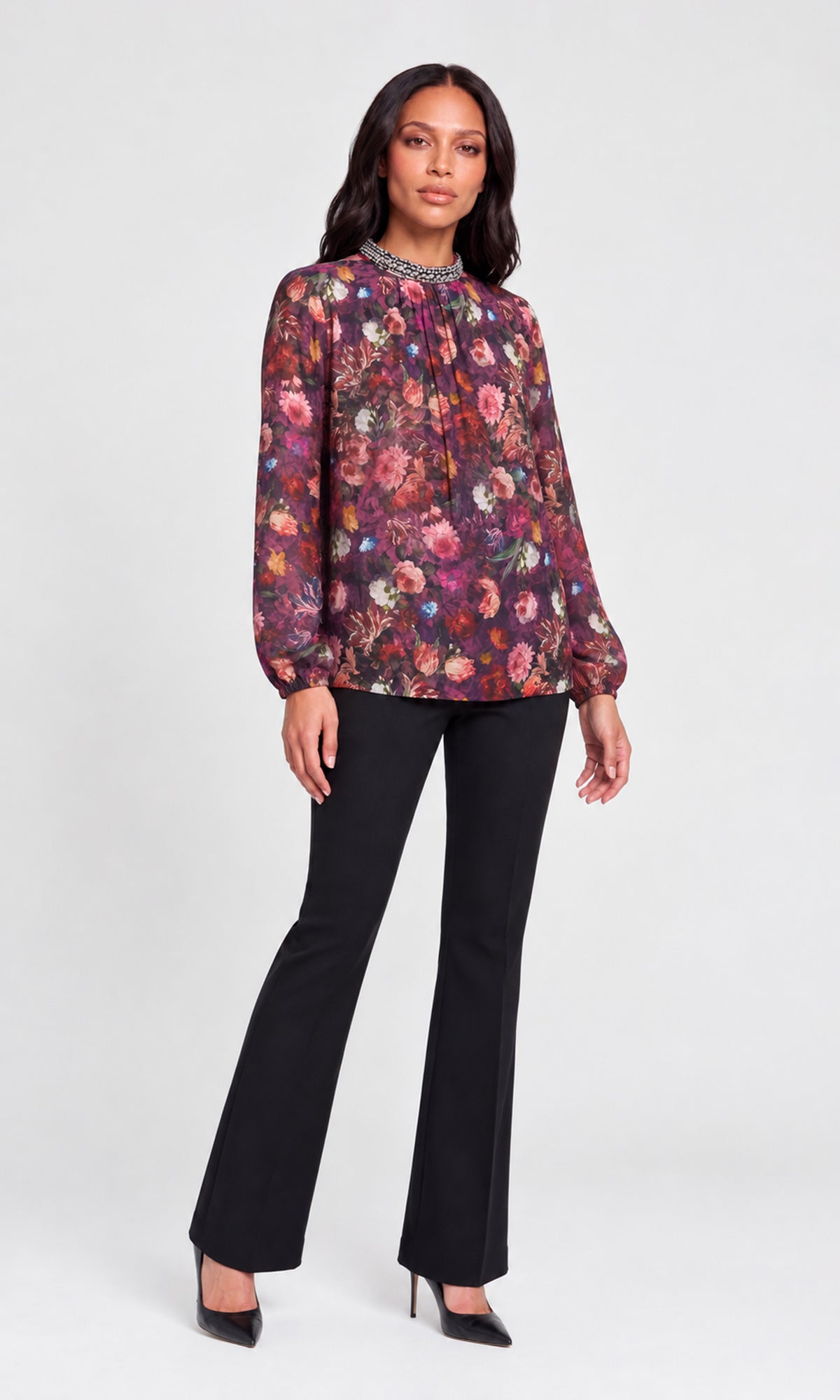 Model wearing a long-sleeve floral blouse with a beaded high neckline, paired with black flared pants and black heels.