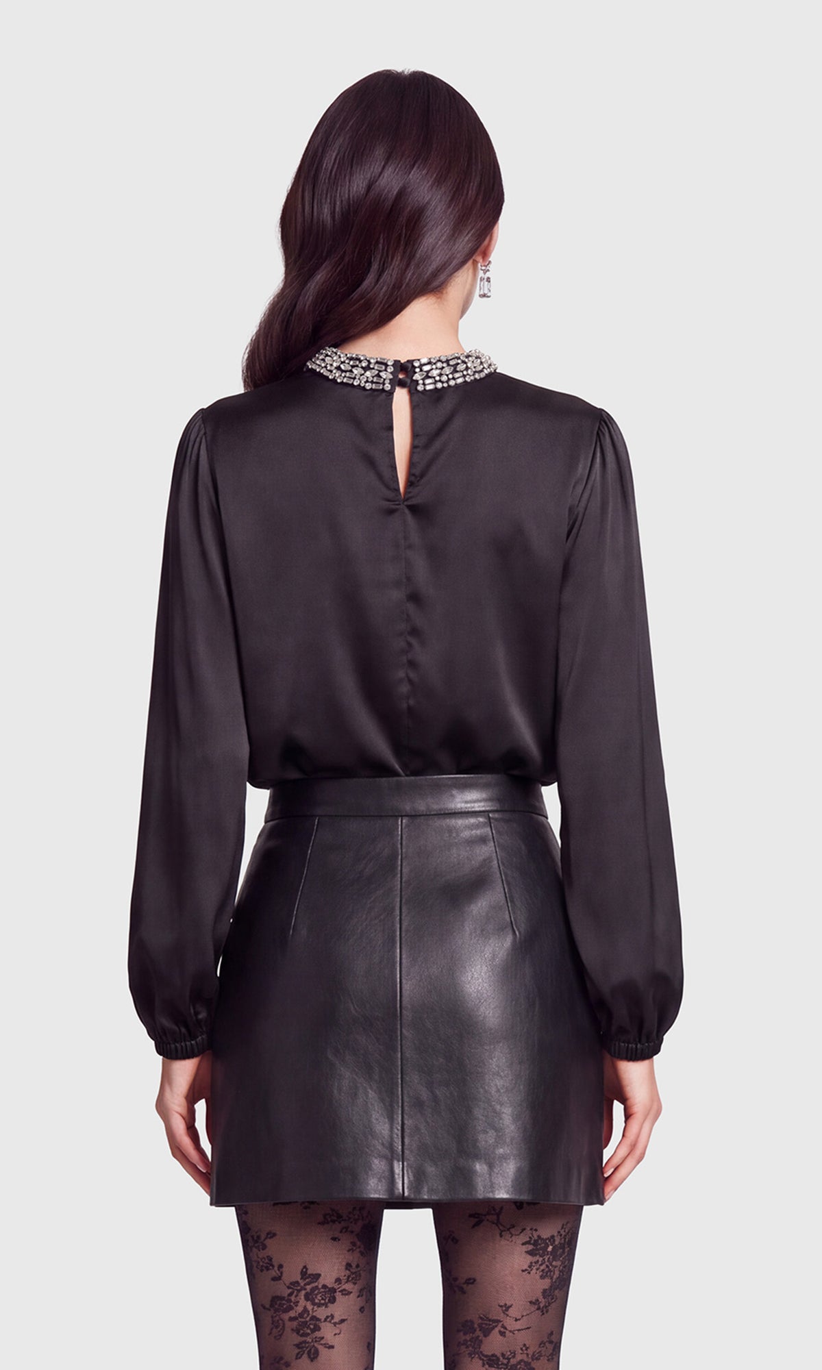 Back view of model wearing a black long-sleeve satin blouse with a jeweled high neckline, paired with a black faux-leather button-front mini skirt and black lace tights.