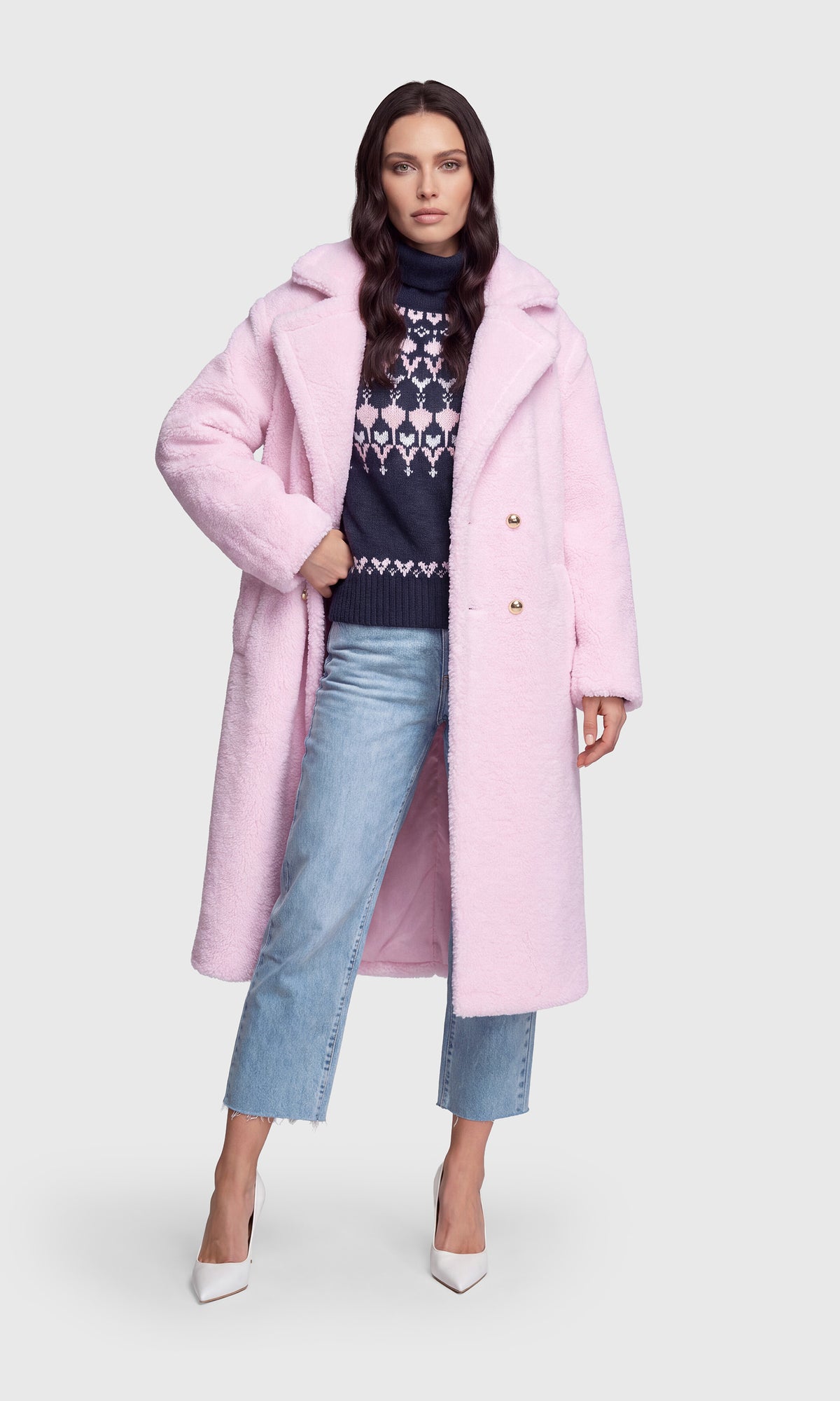 A woman modeling a light pink fuzzy long coat over a navy patterned sweater, light blue jeans, and white pointed heels.