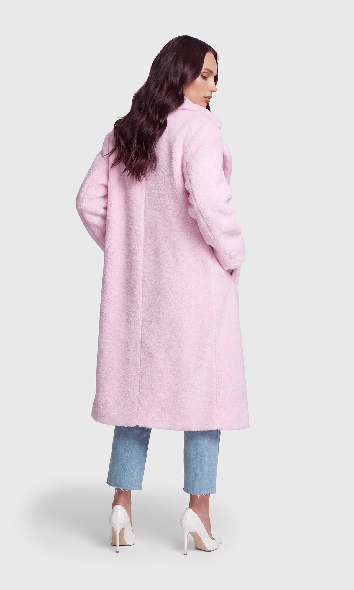 A woman shown from the back wearing a long, light pink fuzzy coat over light blue cropped jeans and white high heels.
