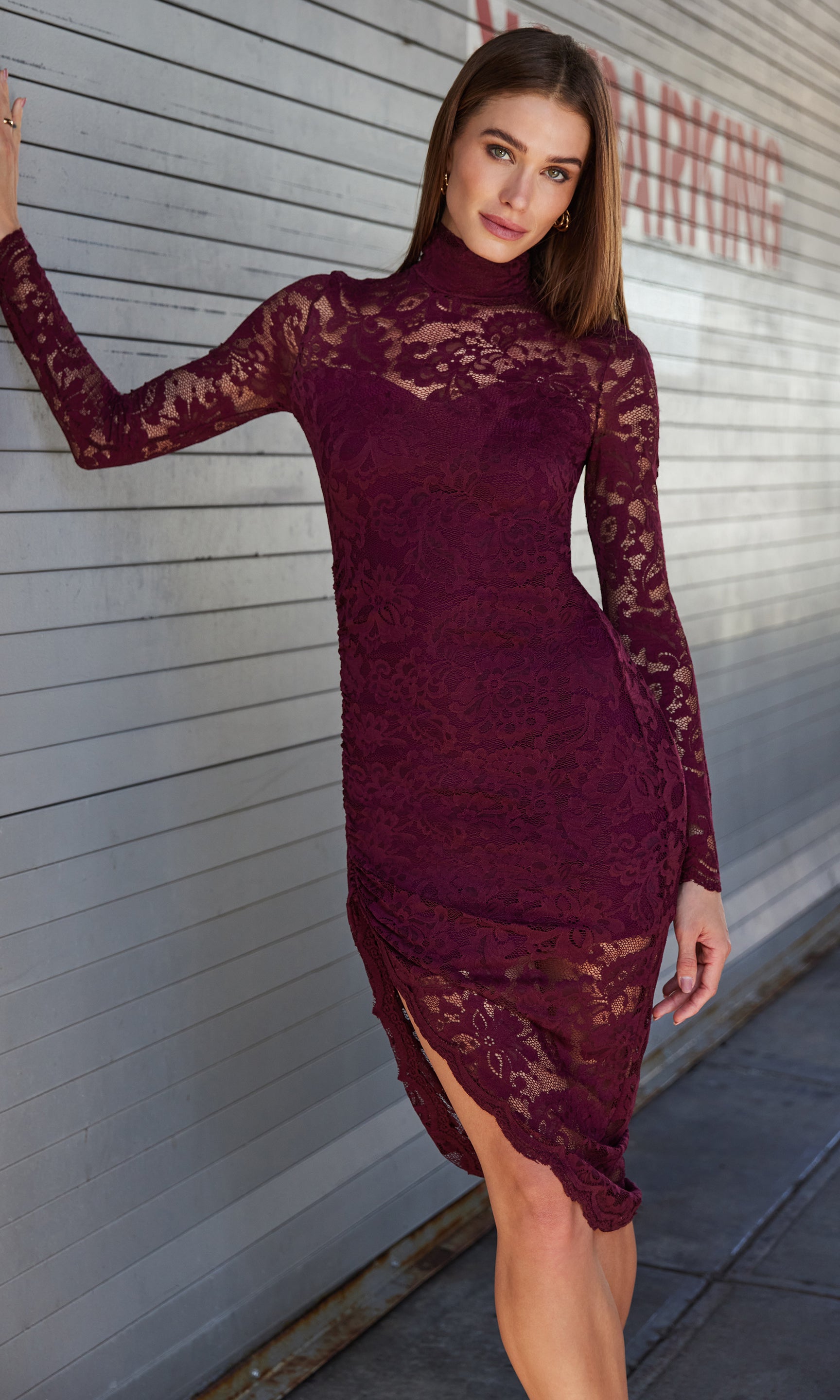 Leaning casually against a wall, she wears the Iria Lace Dress in deep burgundy, featuring sheer lace sleeves and a scalloped hem.