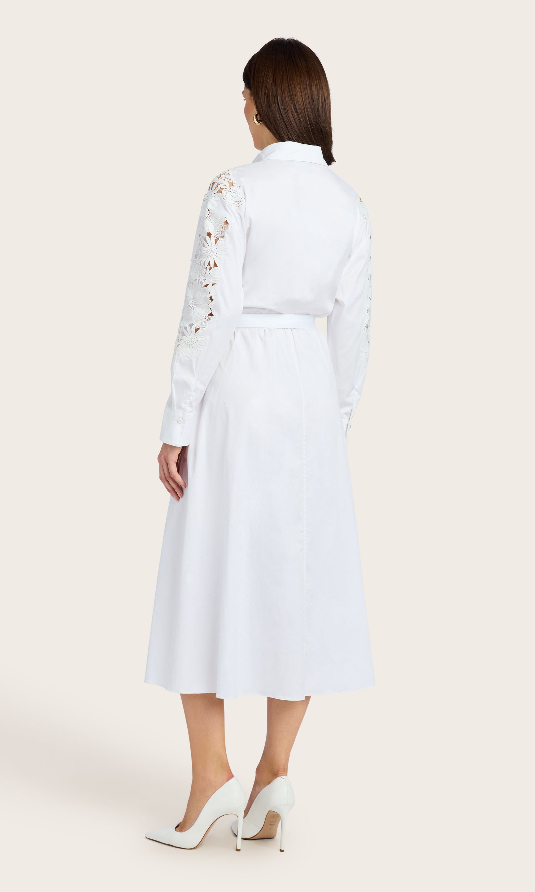 Back view of the model wearing a white button-down lace combo dress with a belted waist and long sleeves, styled with white heels