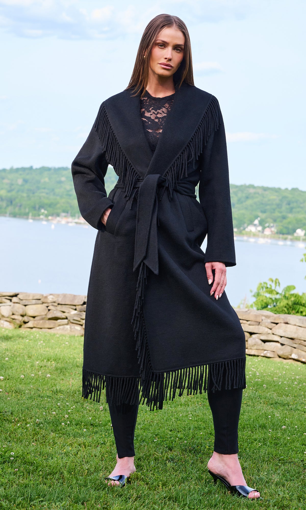 A woman stands outdoors on grass with a lake and hills in the background, wearing a long black fringed wrap coat over a black lace top and black leggings.
