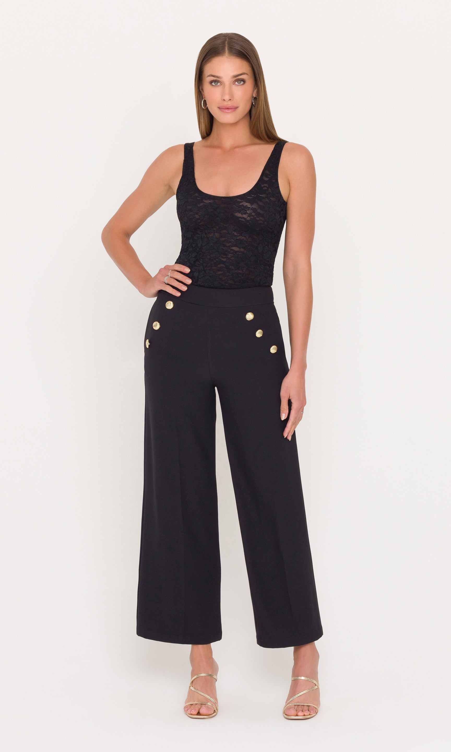 Full body image of woman wearing a black outfit with gold buttons on the pants on a white background
