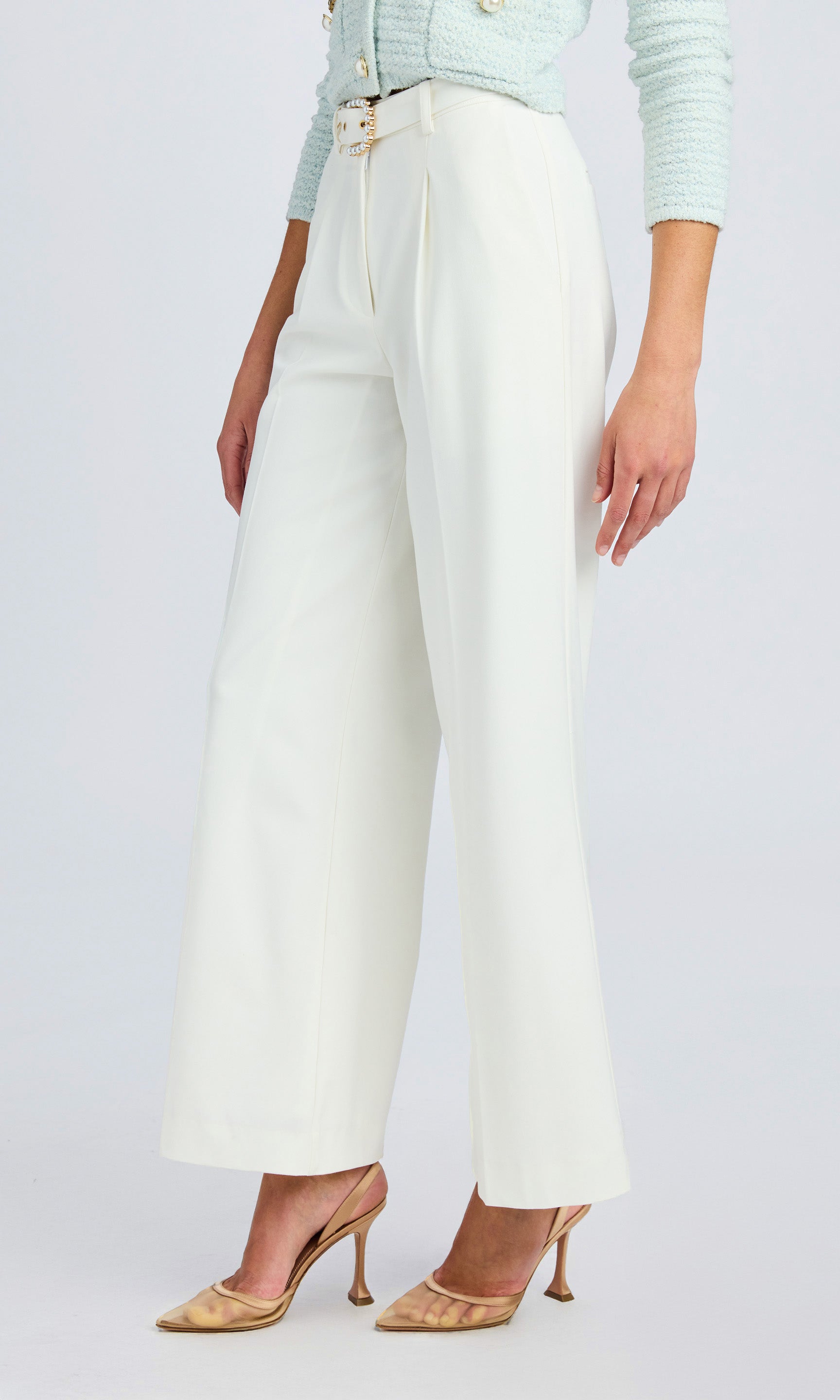 side view of model wearing white crepe pants with faux pearl belt buckle and gold high heels.