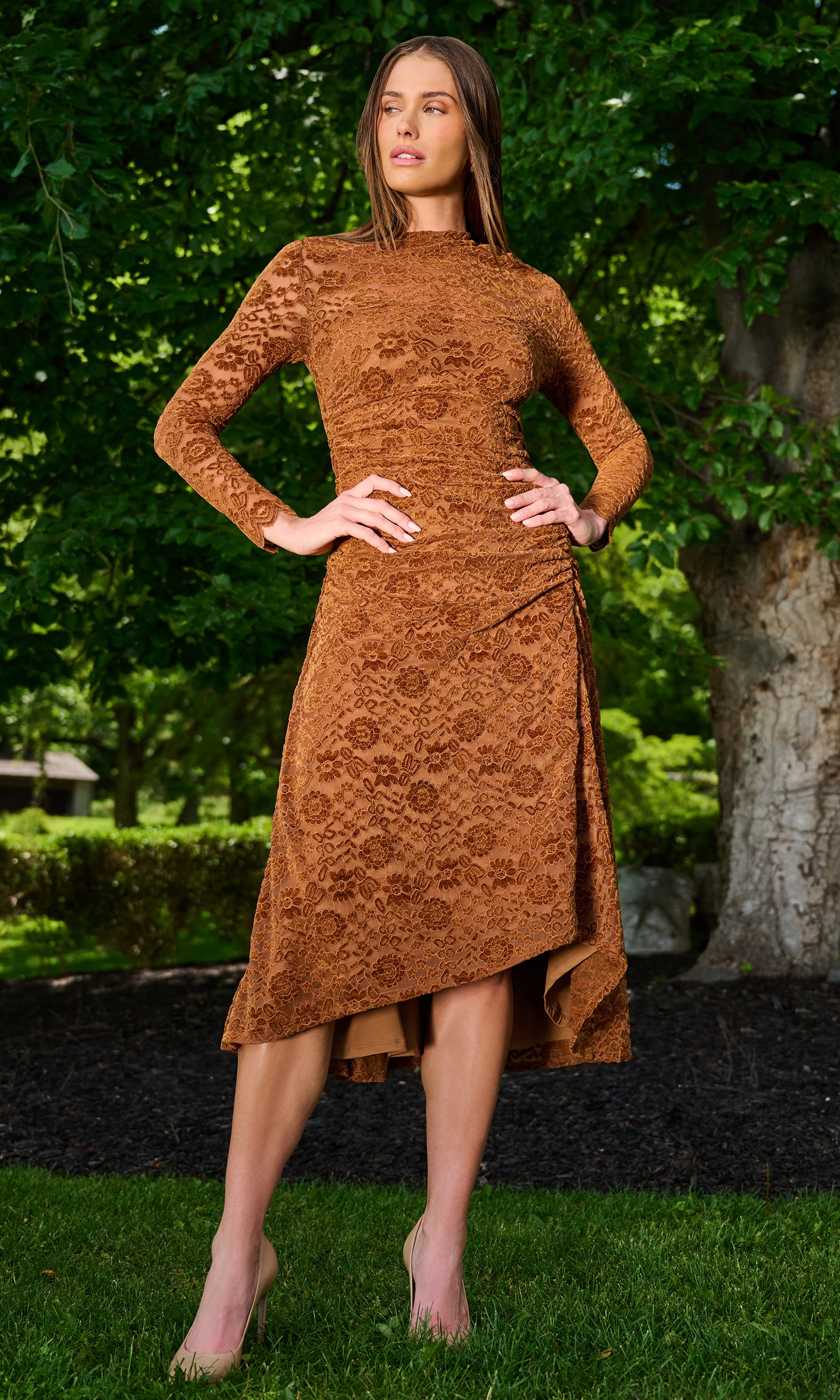 A woman wearing a rust-brown long-sleeve midi dress with a textured floral lace pattern, standing outdoors on a grassy lawn.