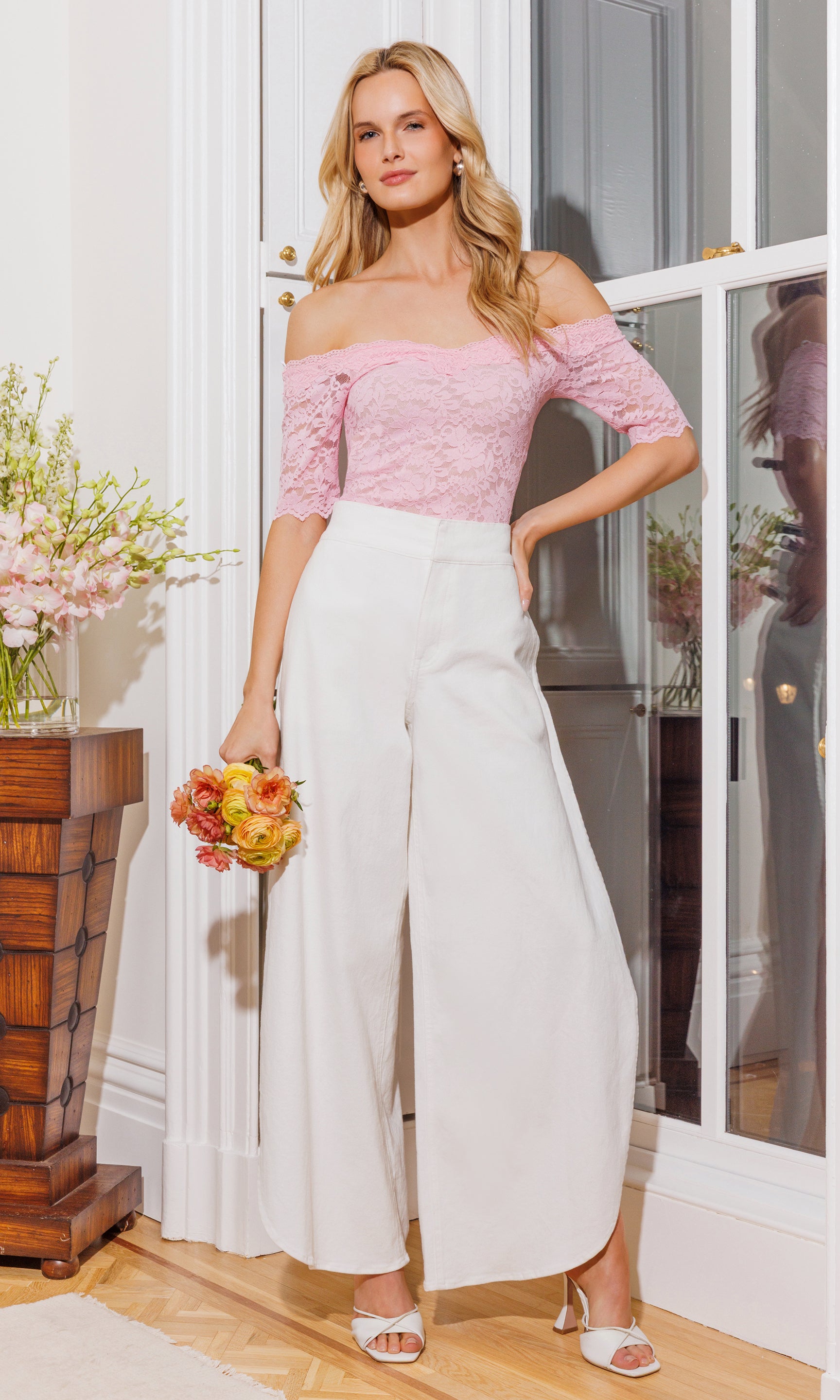 Woman in pink top and white pants holding flowers indoors.