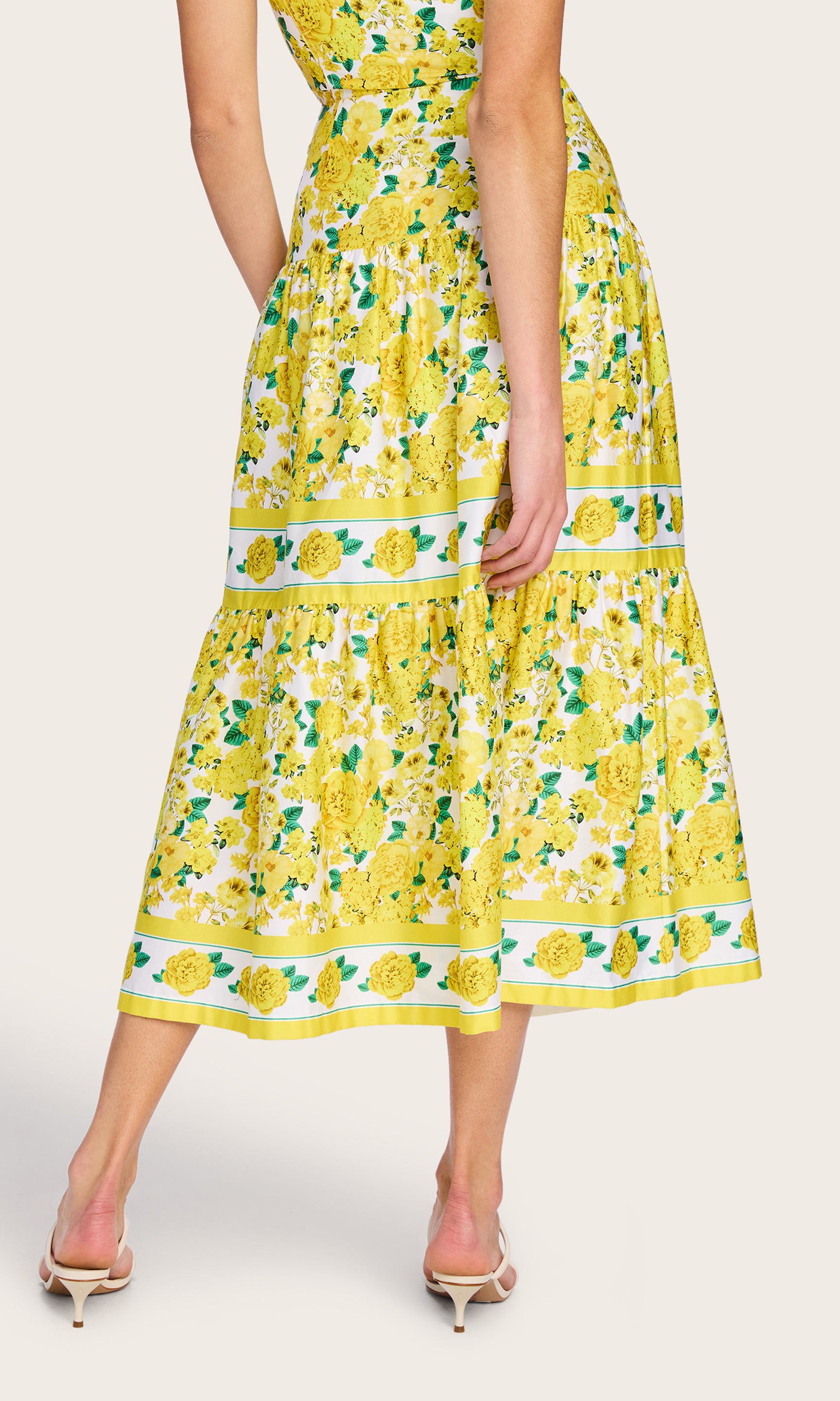 Back view of the model wearing the Mondo Floral Poplin Skirt in a yellow floral print with the matching floral bra top and white heels