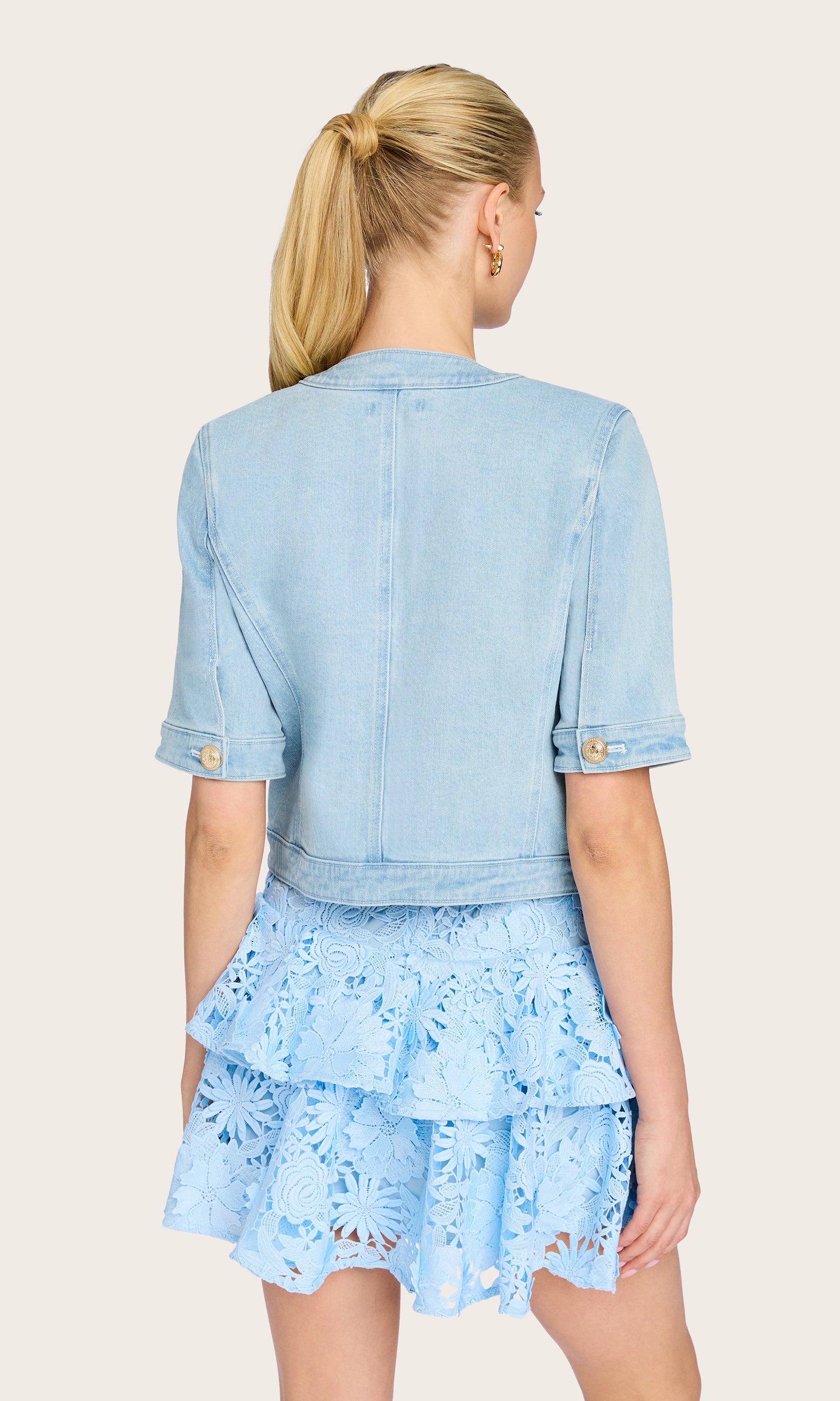 The model turns to reveal the rear view of the Devika Denim Jacket, highlighting the cropped hem and tailored back seams for a structured summer look.