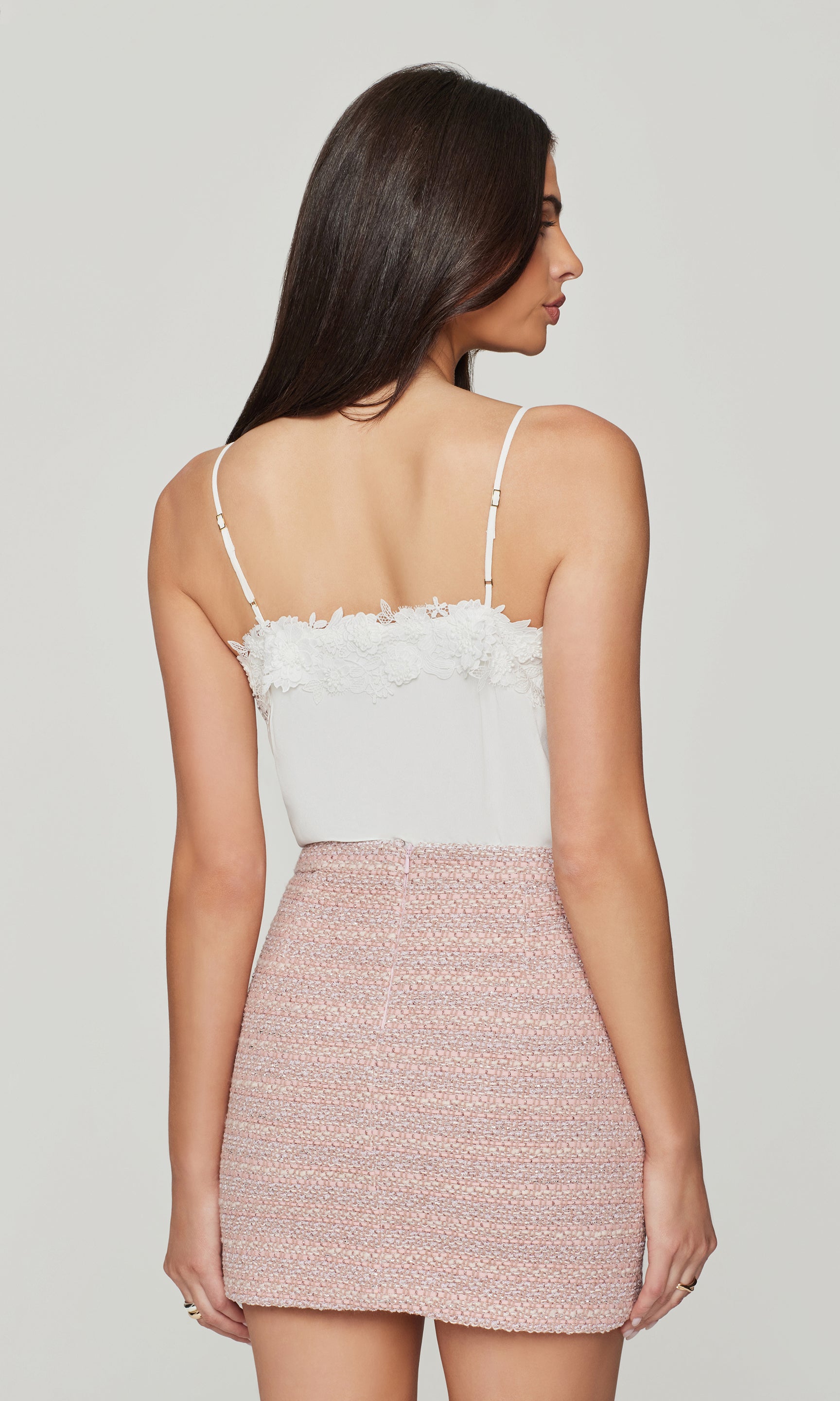 Back view of woman wearing a white cami with lace details and a pink skirt on a plain background