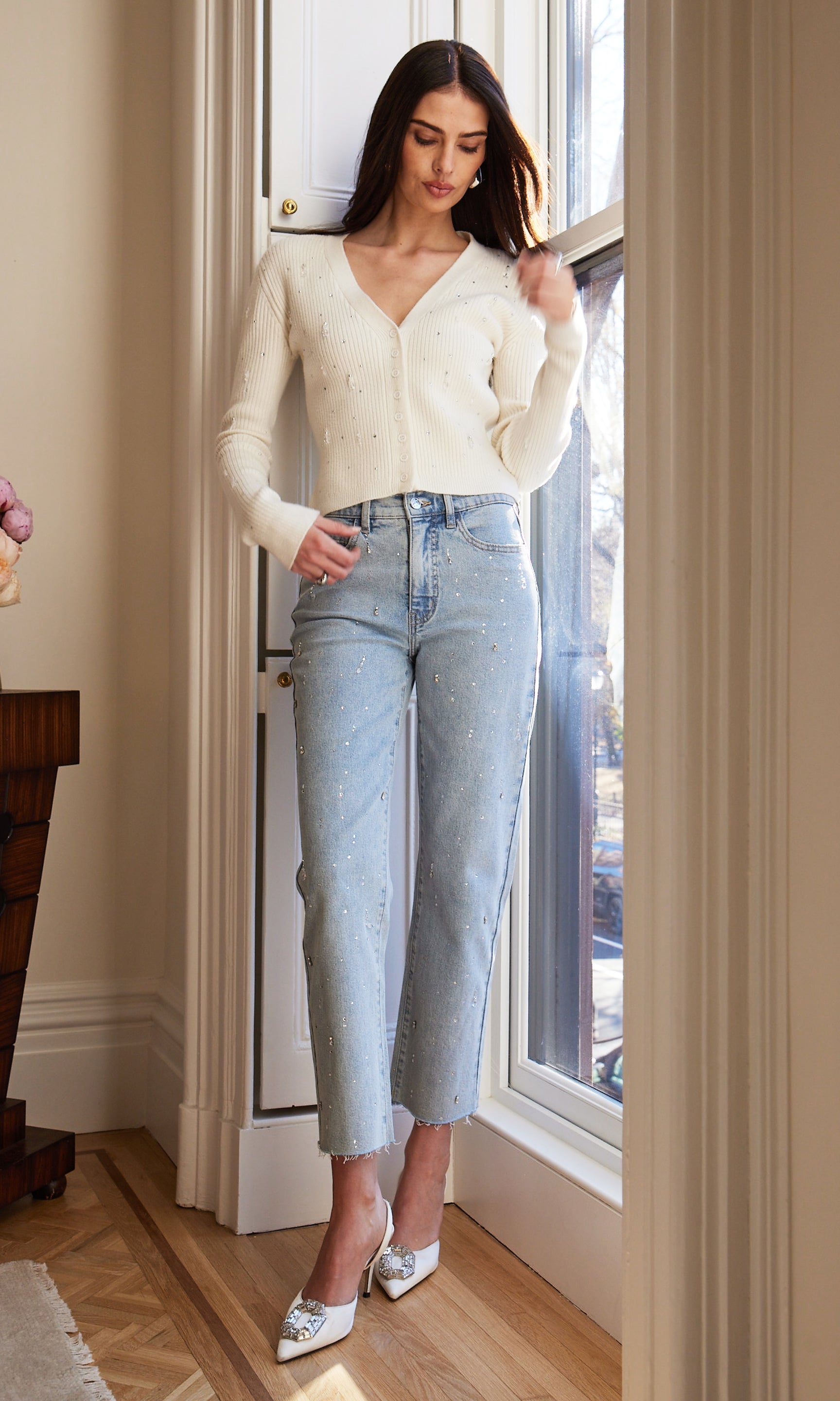 Woman wearing a white cardigan and light blue jeans standing in an indoor setting by a window.