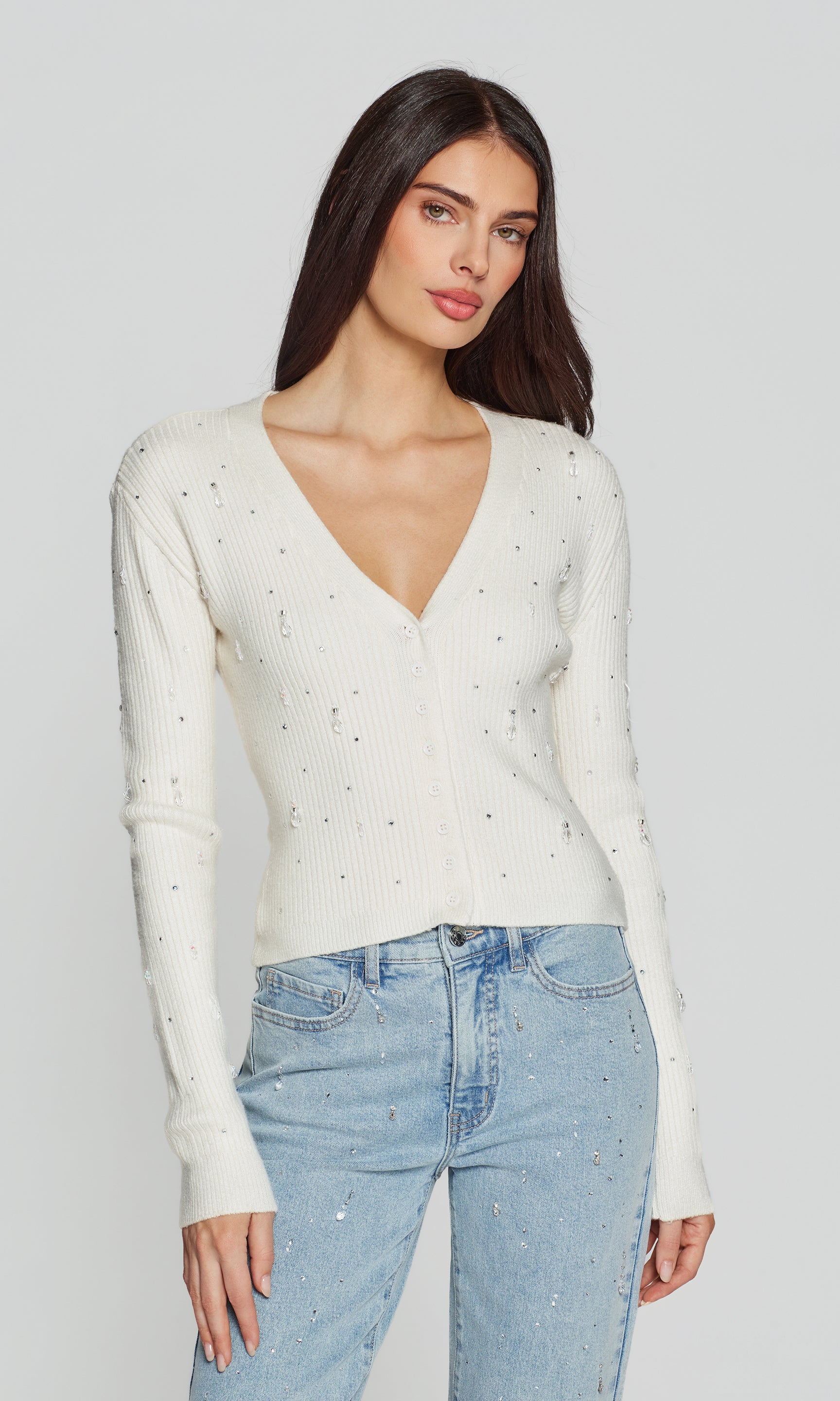 Woman wearing a white sweater with crystal accents and light blue jeans on a plain background