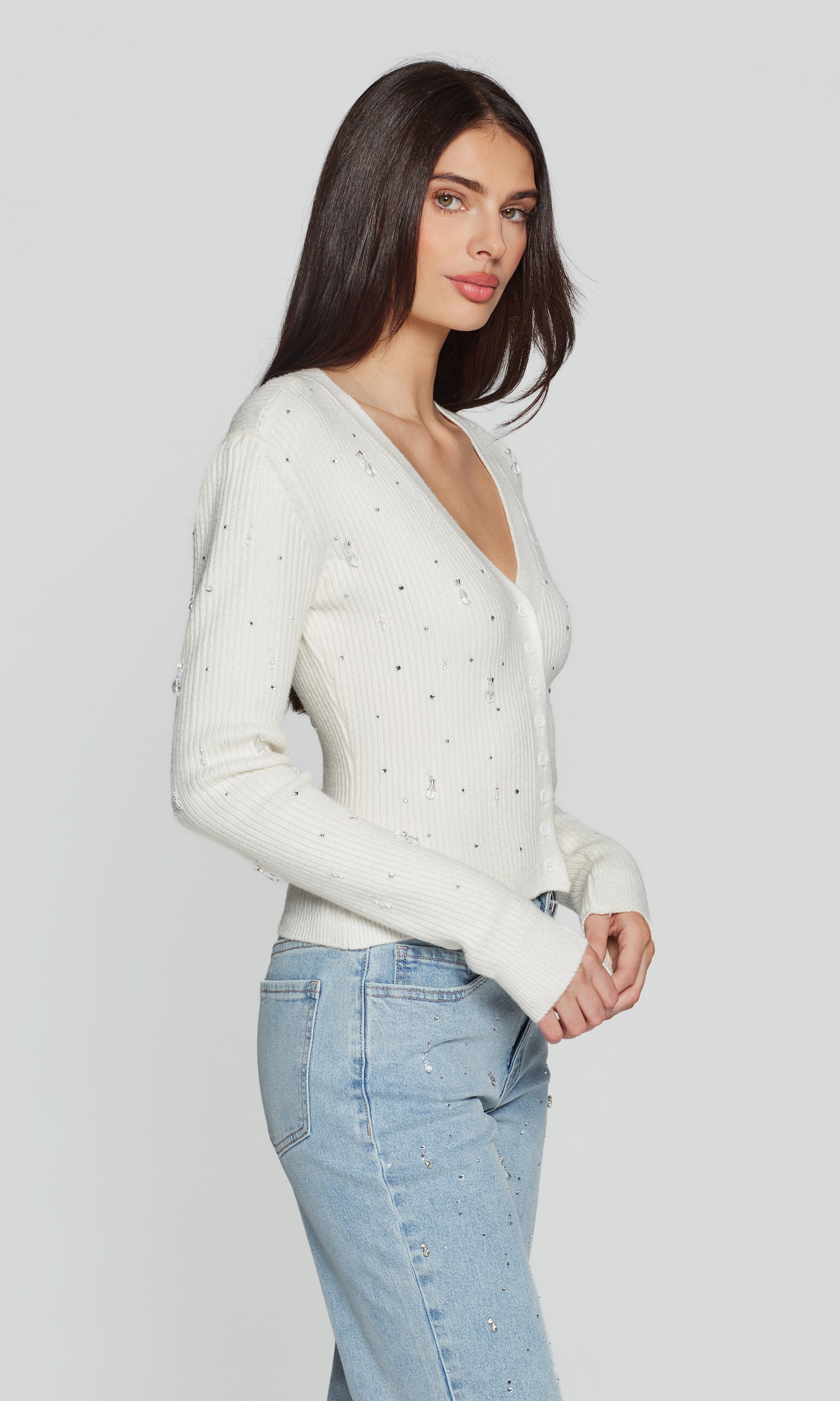 Side view of woman wearing a white sweater with crystal accents and light blue jeans on a plain background