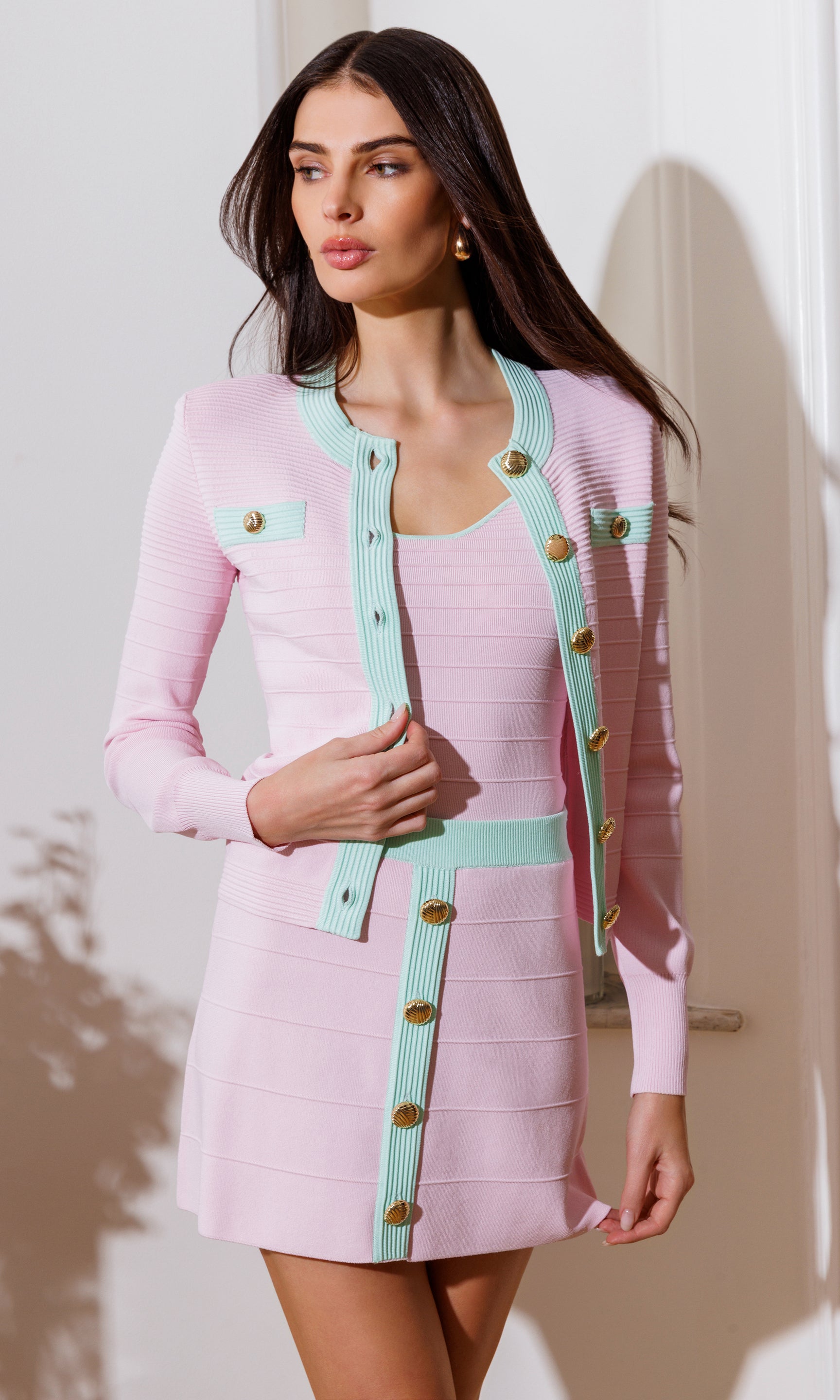 In a three-quarter front view, the model styles the Lilla Bandage Cardigan in pink with the coordinating mini skirt, showcasing the cropped fit and contrast trim.
