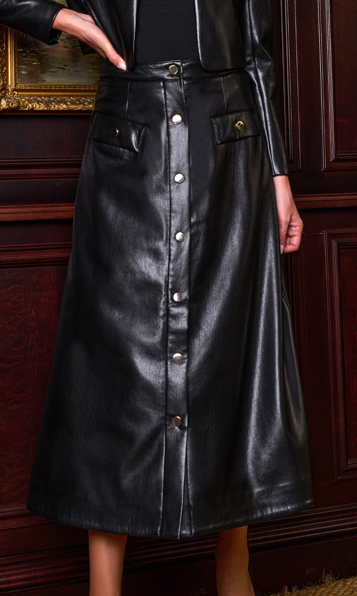 A black faux leather midi skirt with a button-down front and flap pockets, shown on a model in front of a dark wood paneled wall.