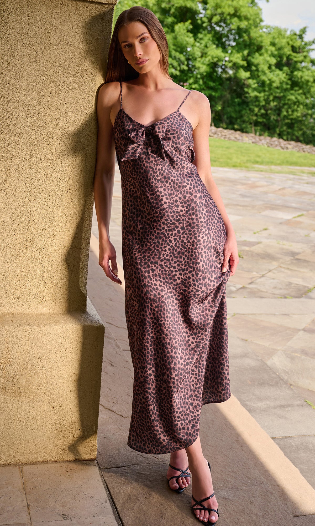 A woman wearing a leopard-print spaghetti-strap maxi dress with a bow at the bust, standing outdoors next to a beige stucco column on a stone patio.
