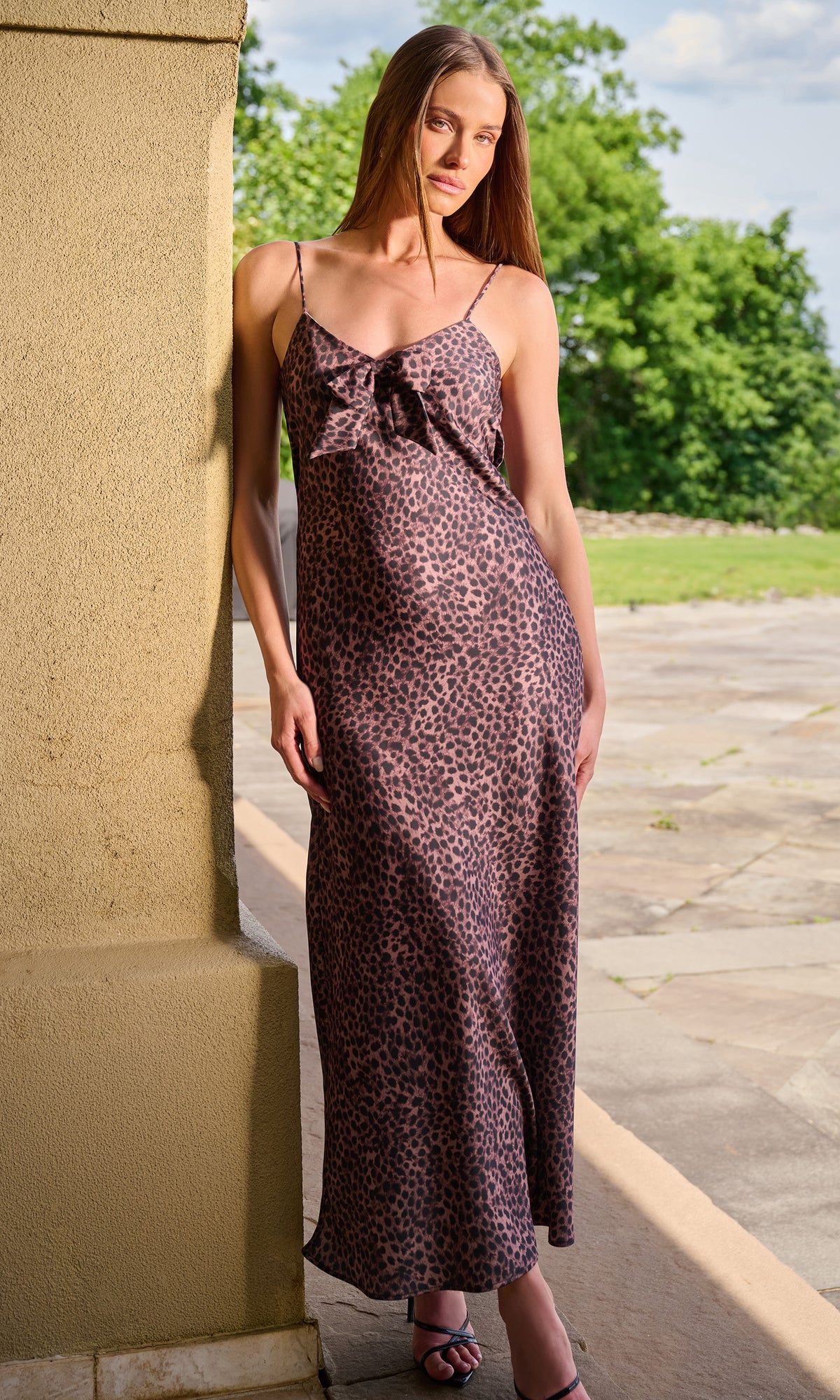 A woman wearing a sleeveless leopard-print maxi dress with spaghetti straps and a large bow detail at the bust, standing outdoors.