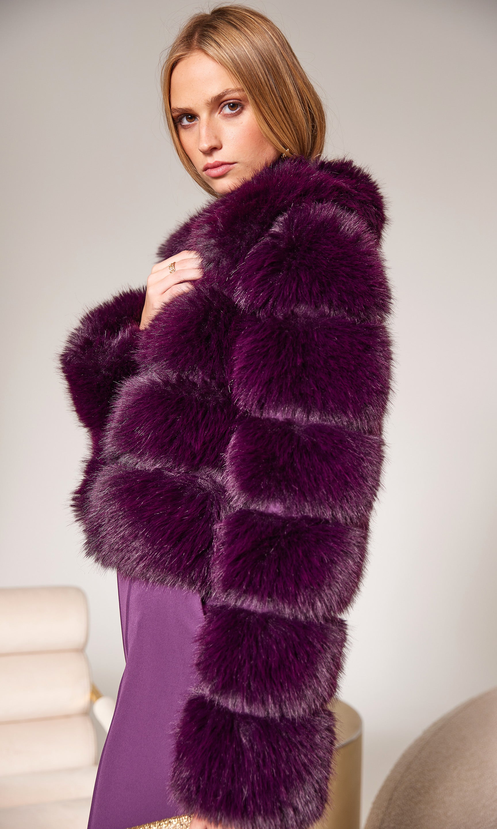 Woman wearing a long, dark purple fur coat against a plain background