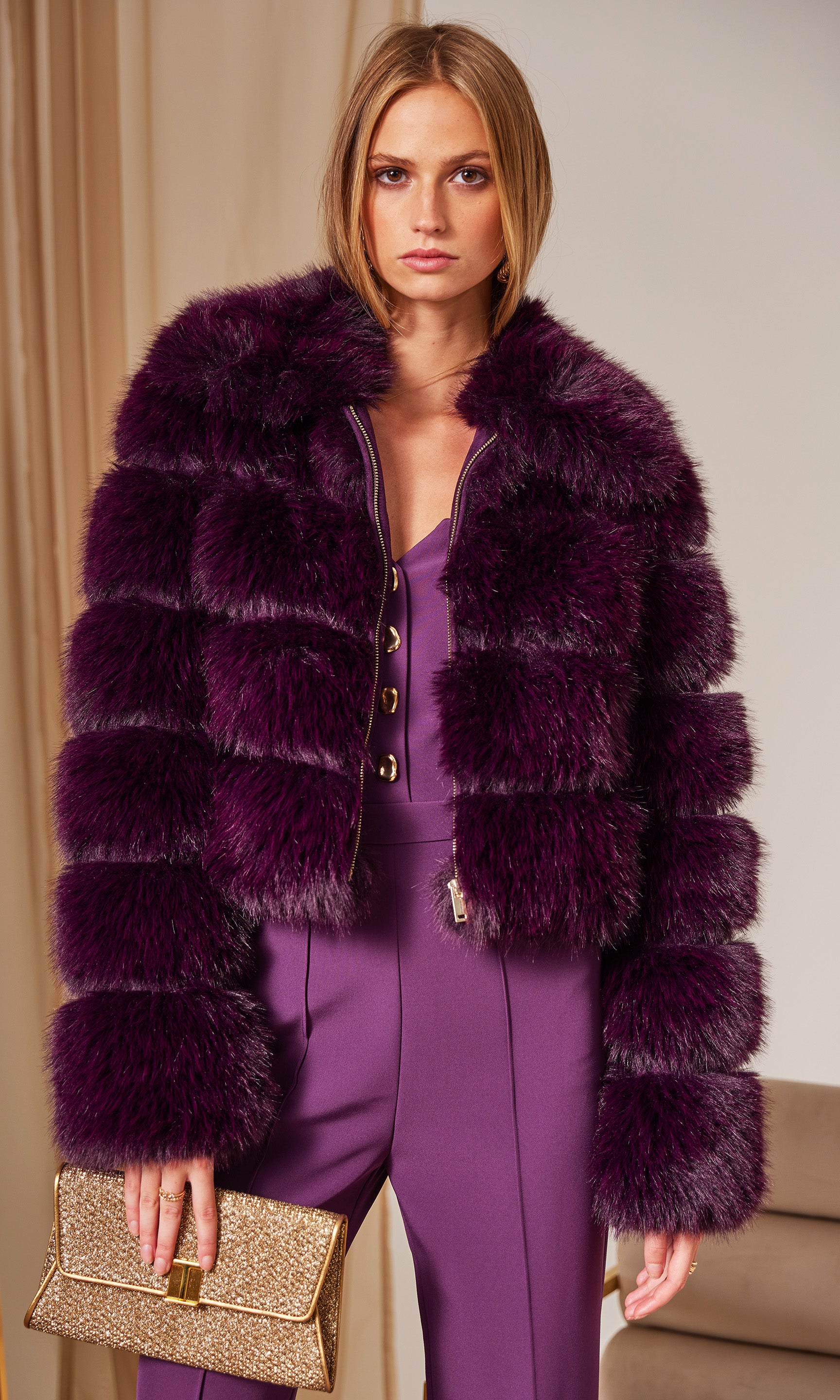 Woman wearing a purple fur coat over a matching outfit, holding a gold clutch.