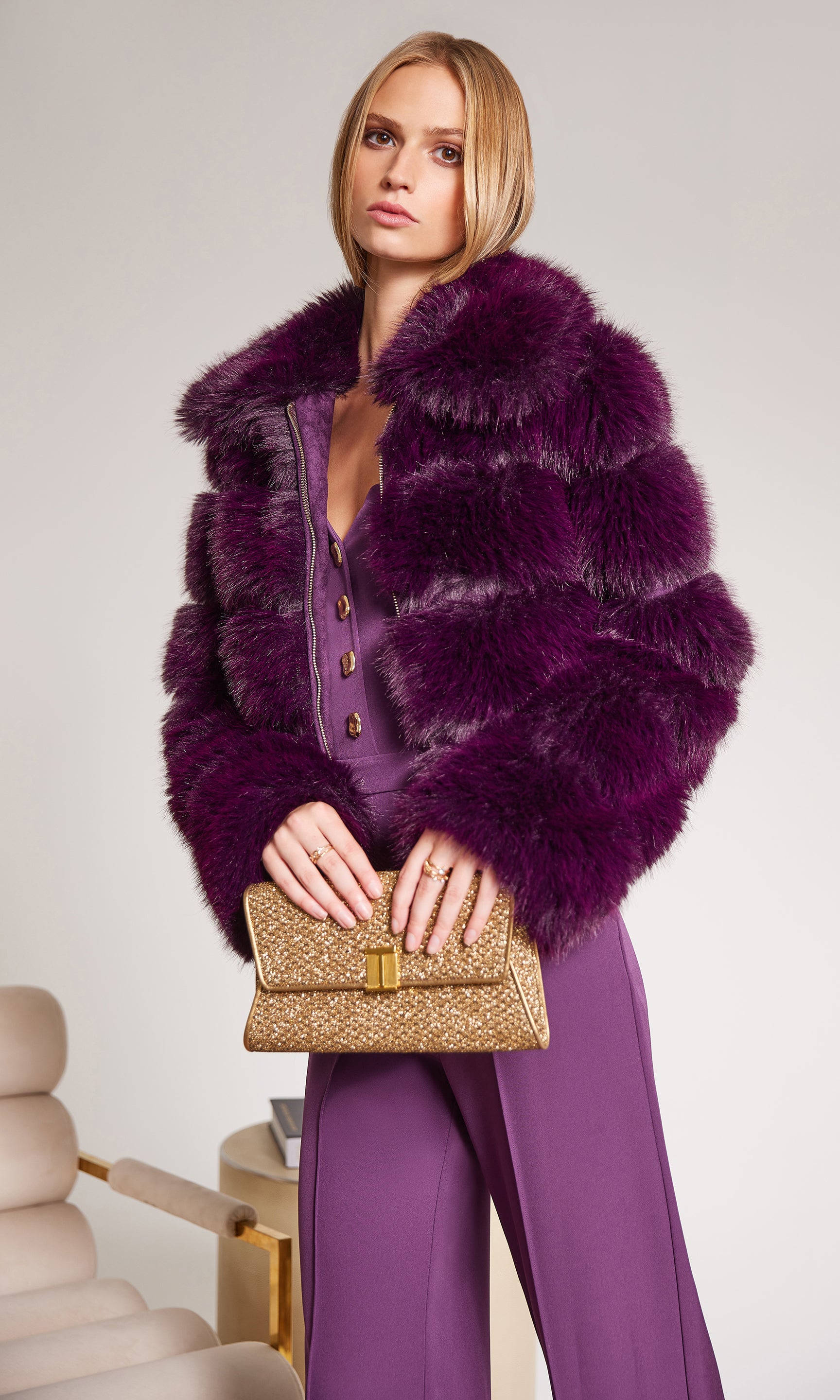 Woman wearing a purple fur coat and matching pants, holding a gold clutch.