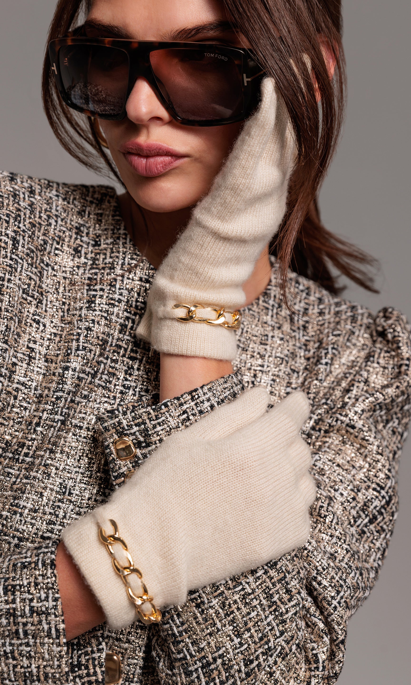 Person wearing sunglasses, white gloves with gold chains, and a textured coat on a gray background