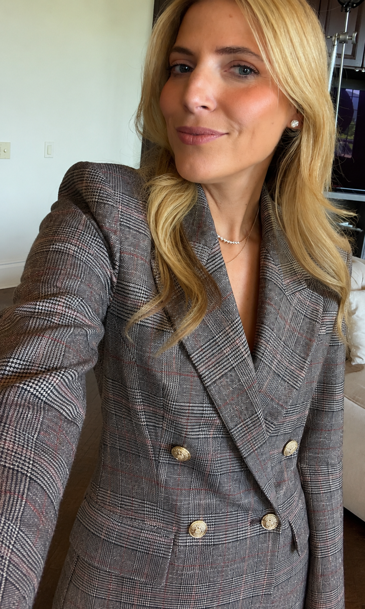 A woman wearing a gray double-breasted plaid blazer with gold buttons, taking a mirror selfie in an indoor setting.
