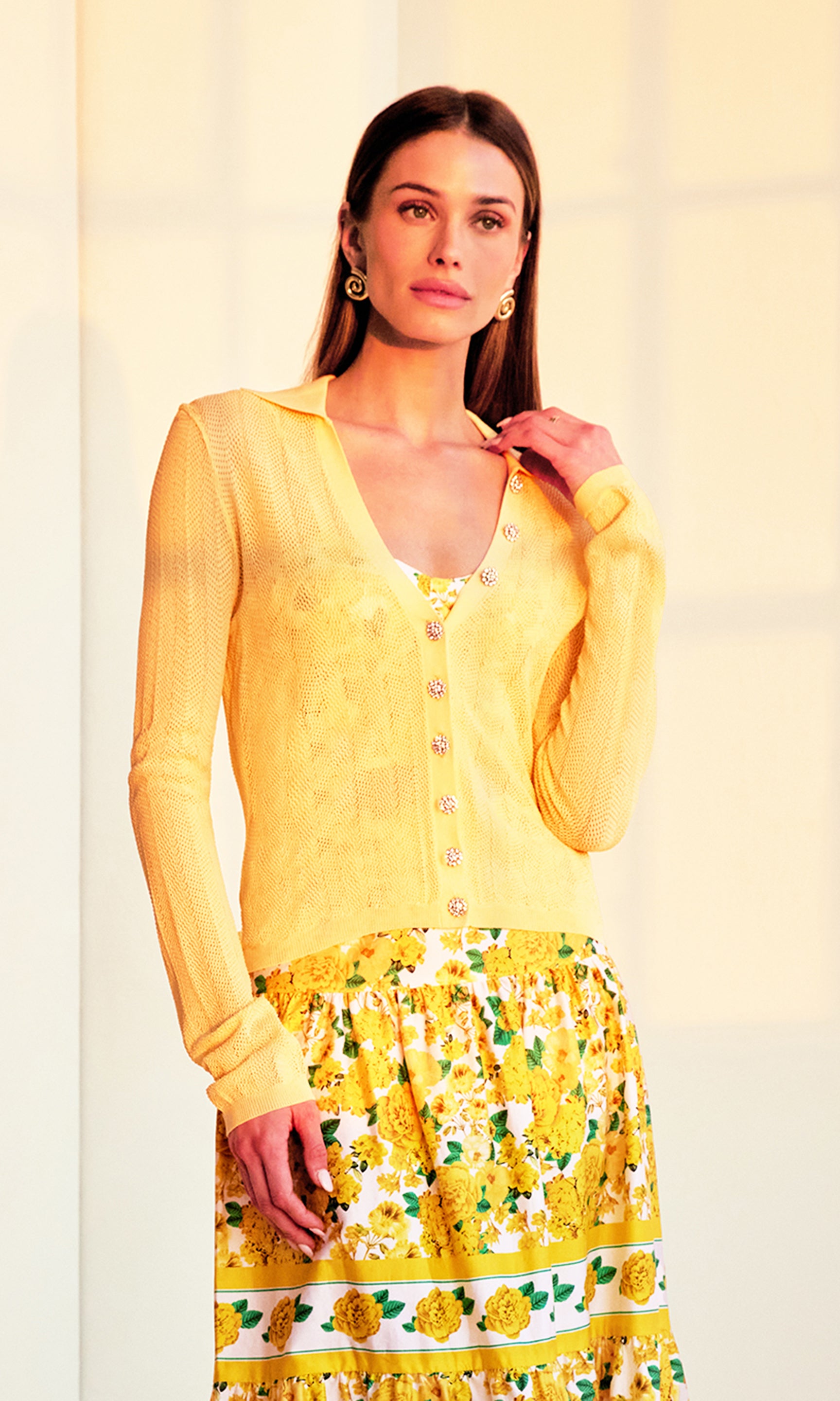 front crop image of model in a sunset-lit room posed with her hand subtly touching her own shoulder wearing the Megan Pointelle Cardigan in pale yellow showing the crystal buttons over a yellow floral top and skirt.