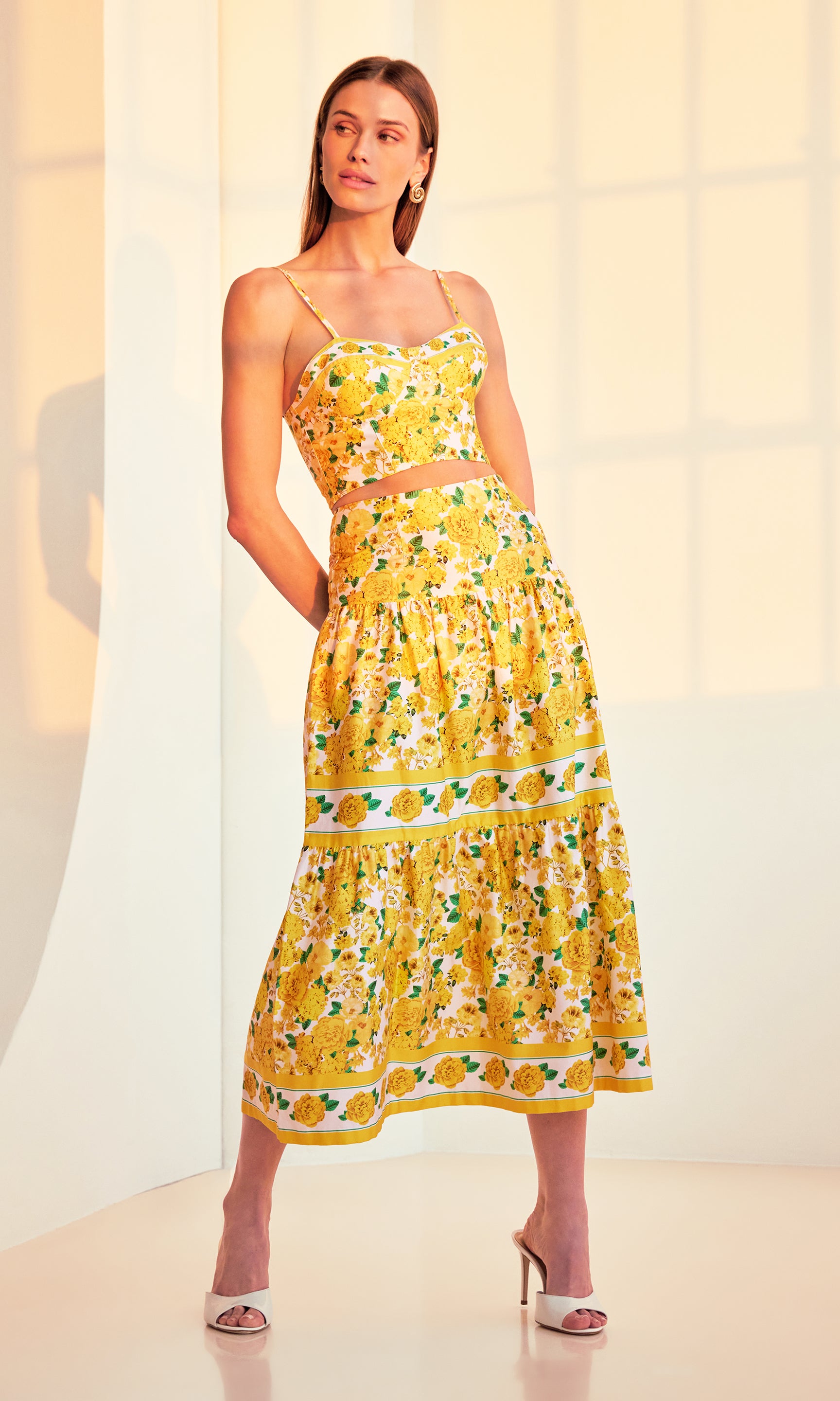 full body image of model posing in a sunset-lit room with her hands behind her back wearing a yellow floral top with the matching Mondo Floral Poplin Skirt, paired with white heels.