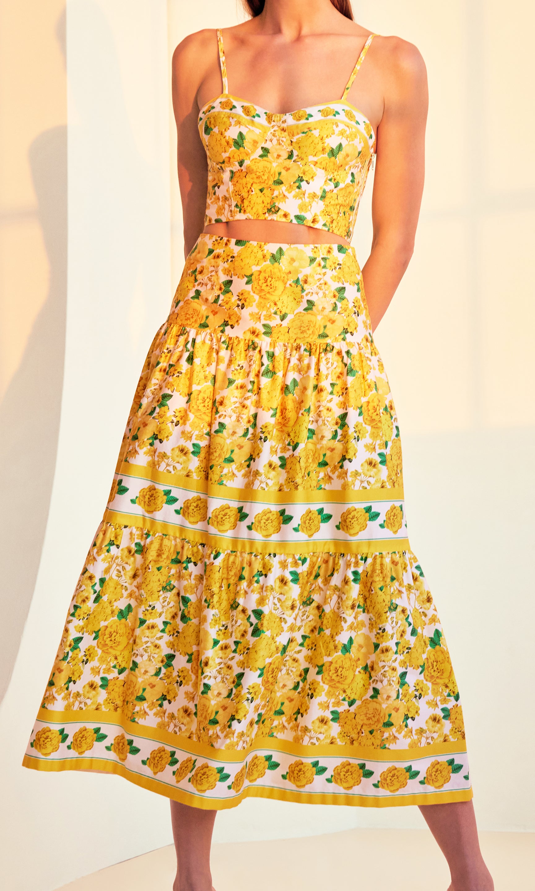 Zoomed in image of model posing in a sunset-lit room with her hands behind her back wearing a yellow floral top with the matching Mondo Floral Poplin Skirt showcasing w=the beautiful pattern all over and tiered structure of the skirt.