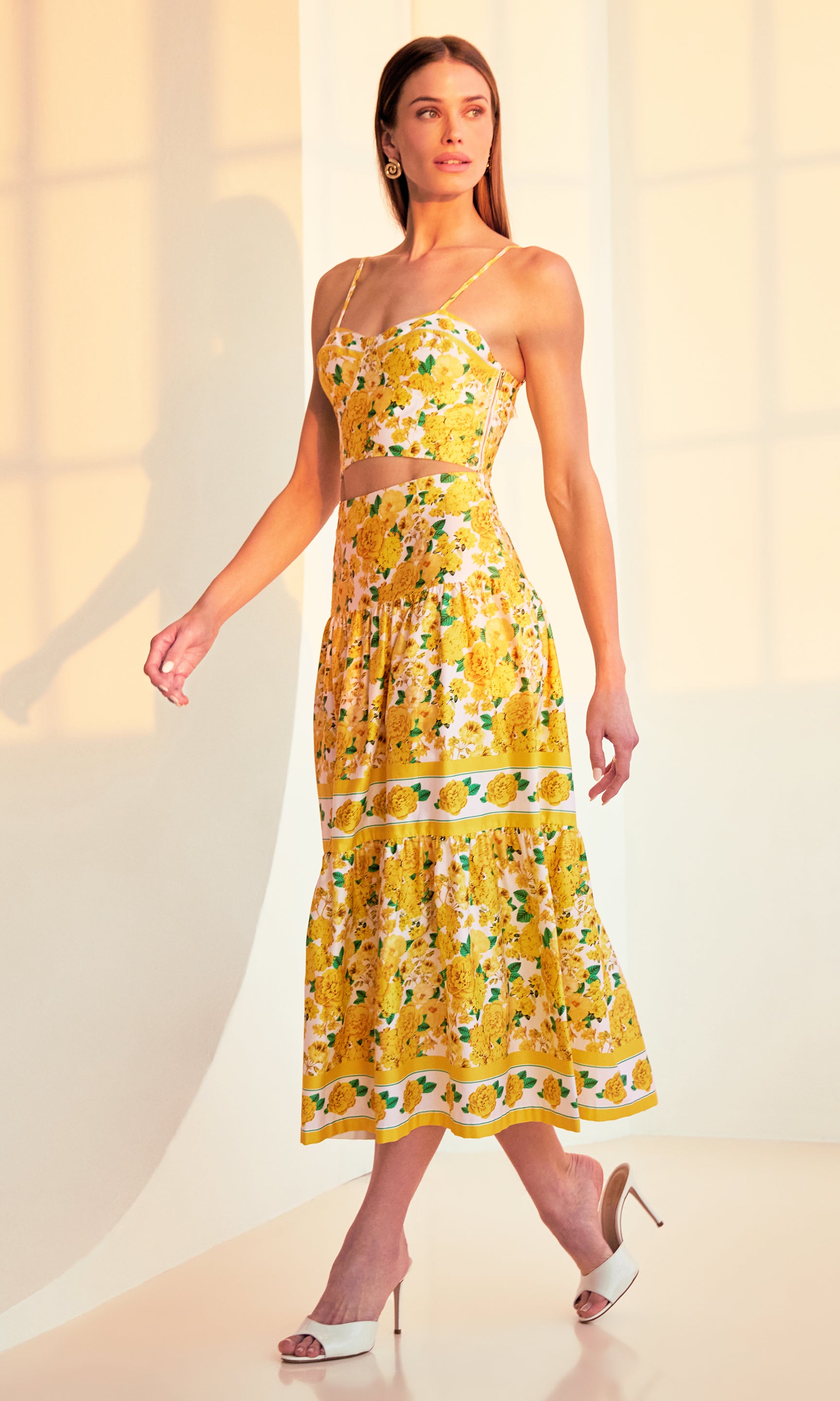 side view full body image of model walking in a sunset-lit room with her hands behind her back wearing a yellow floral top with the matching Mondo Floral Poplin Skirt, paired with white heels.