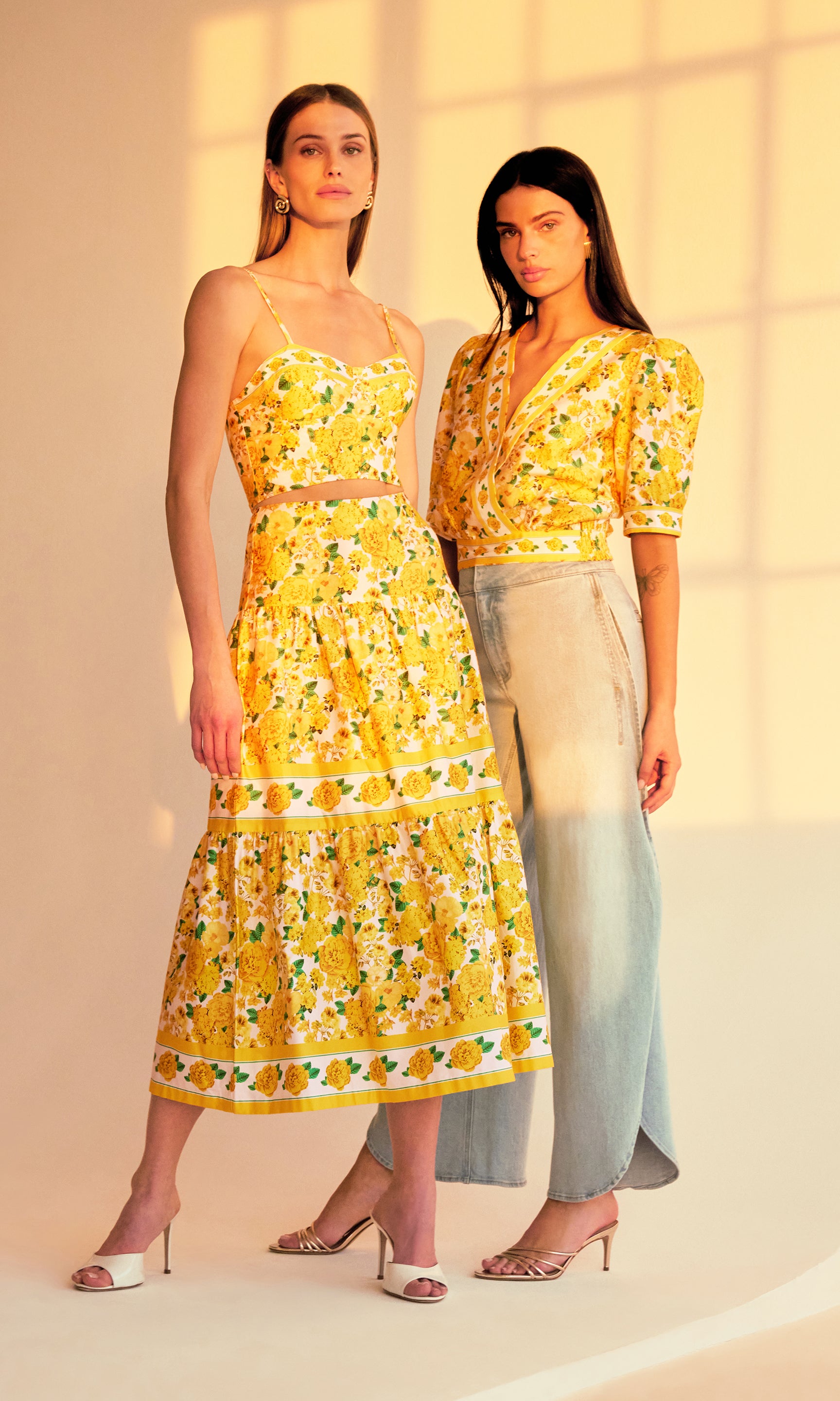 image of two models posing in a sunset-lit room both wearing yellow floral styles, on the left she is wearing a bra cami top and the matching Mondo floral poplin skirt in the yellow floral print, on the right she is wearing a blouse in the same yellow floral pattern with blue jeans.