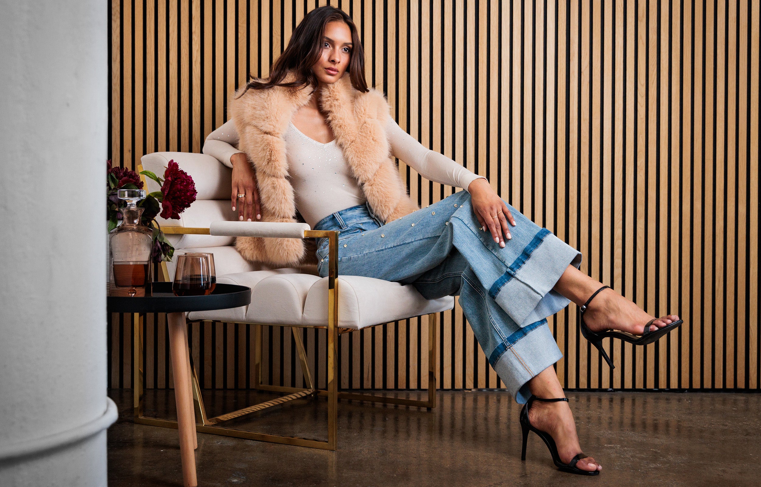 Woman sitting on a chair wearing a fur vest and blue crystal embellished jeans in an indoor setting.
