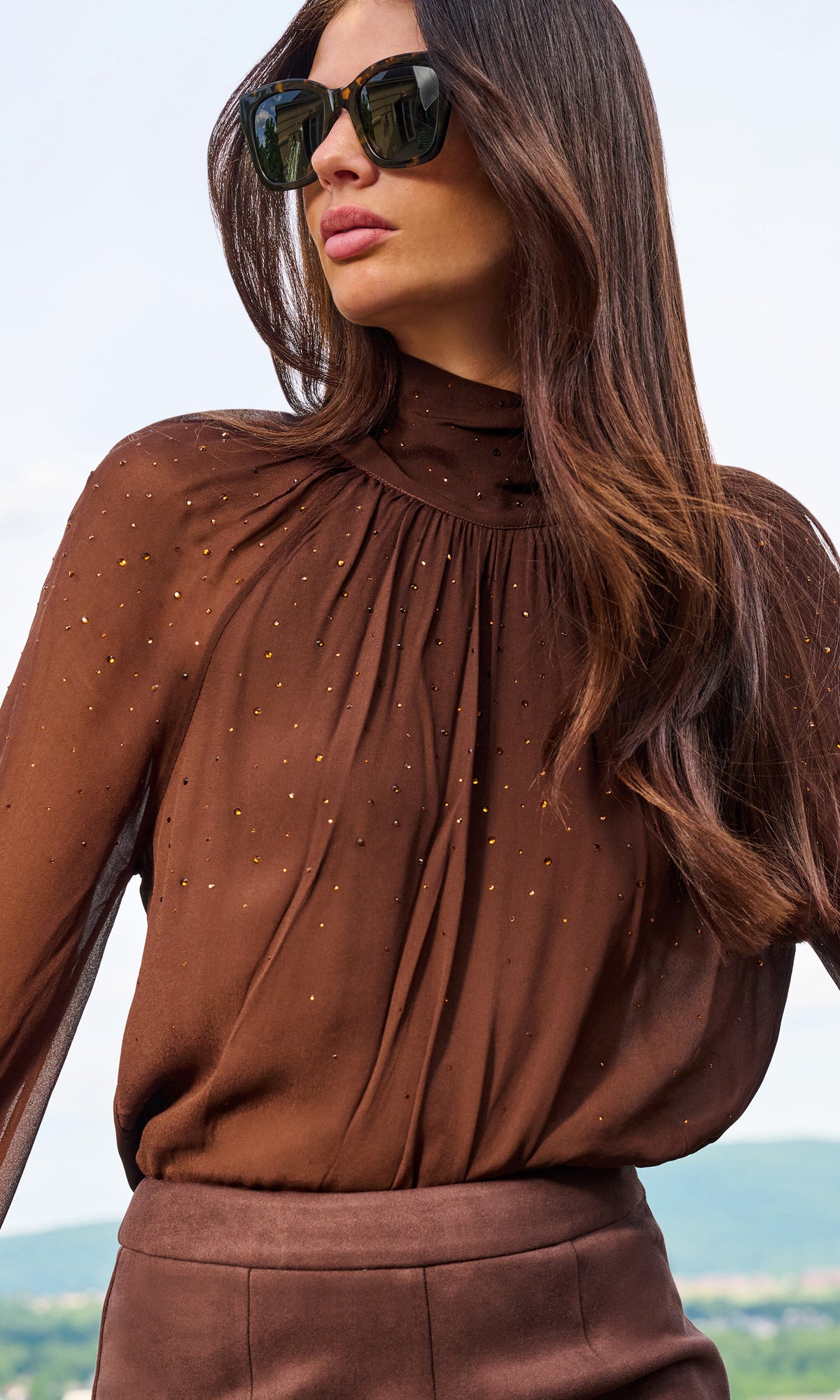 A woman wearing a brown sheer long-sleeve blouse with gold sequin embellishments and a high gathered neckline, paired with brown bottoms and dark sunglasses.