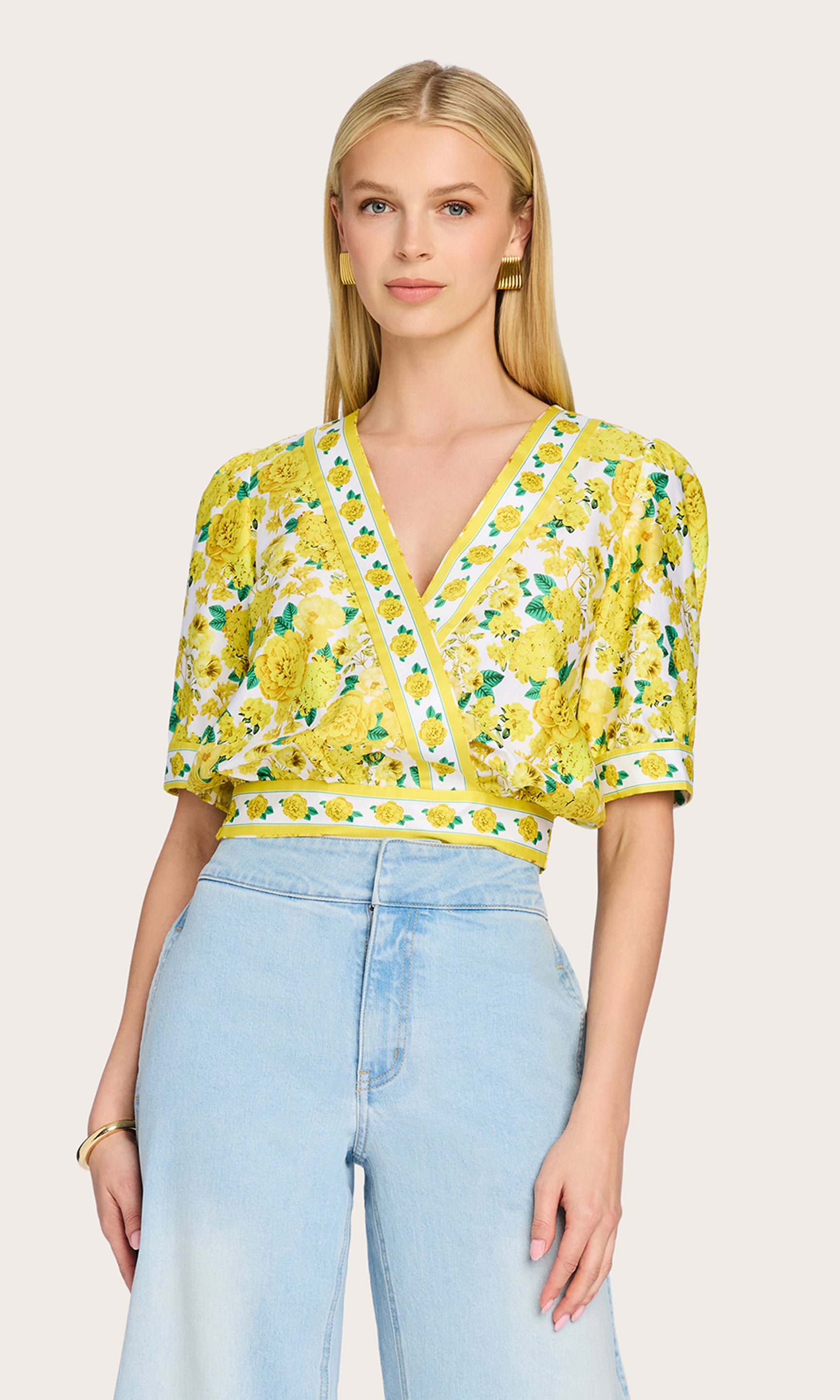 Front view image of the model wearing the Rema Floral Poplin Blouse in a yellow floral print, highlighting the wrap silhouette and puff sleeves styled with blue high-rise jeans.