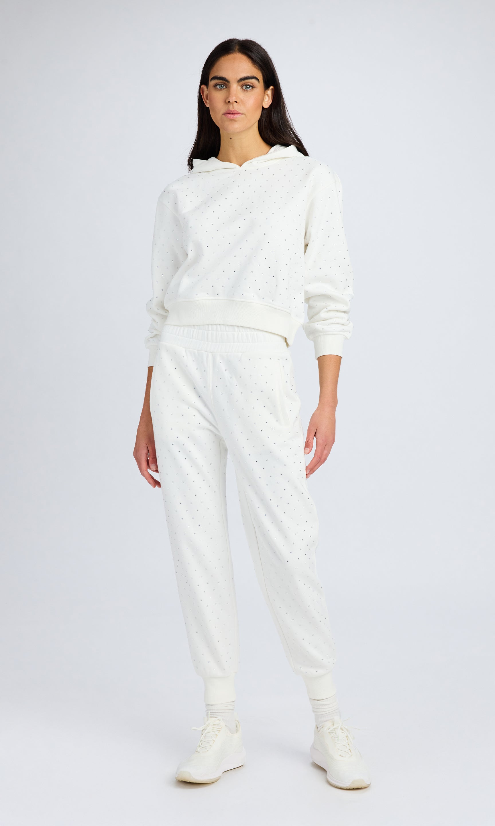 Model with dark hair stands wearing a white crystal sweatshirt and matching sweatpants.