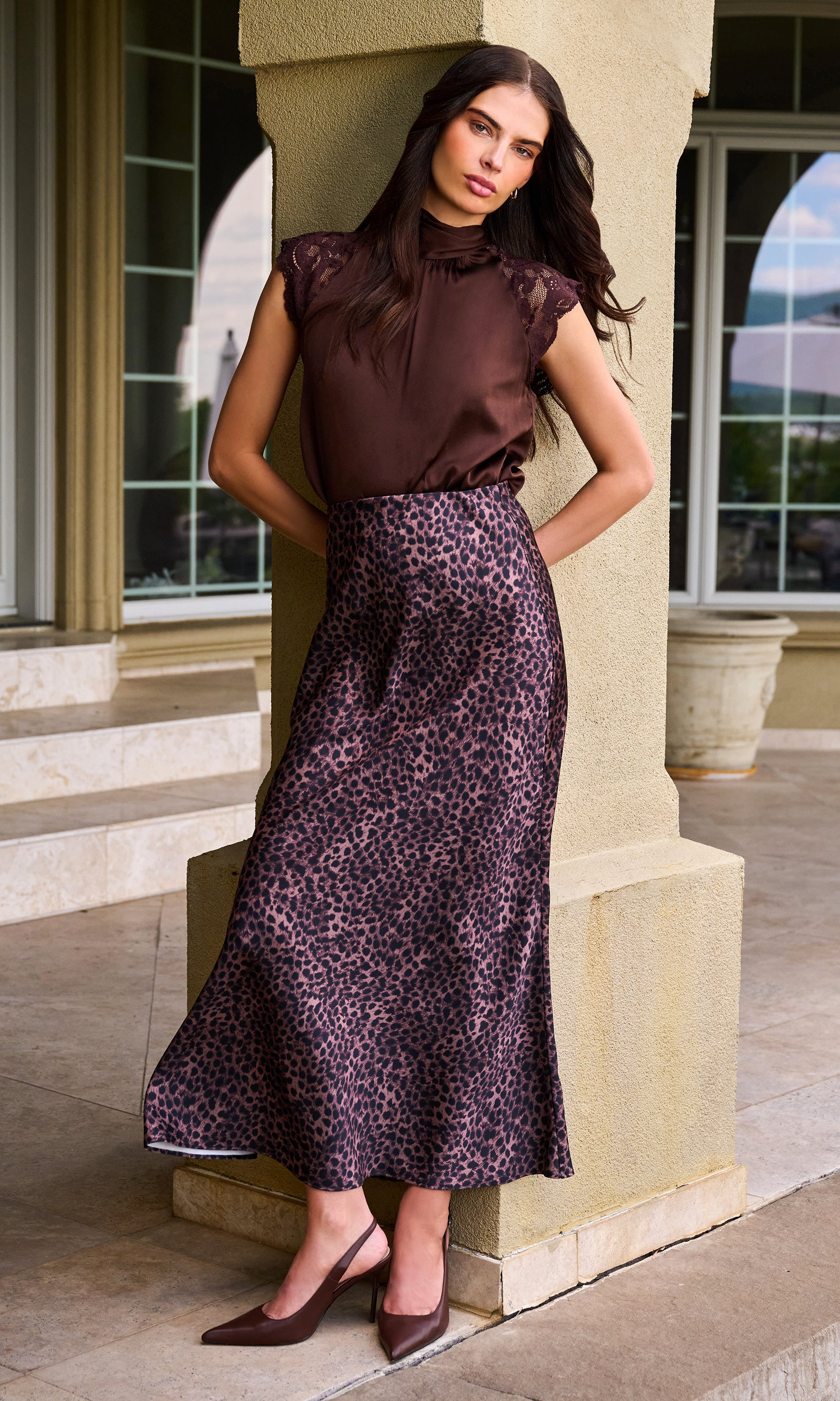 Model wearing a dark brown satin halter-top and a matching leopard-print midi skirt, standing in front of a beige stone column.