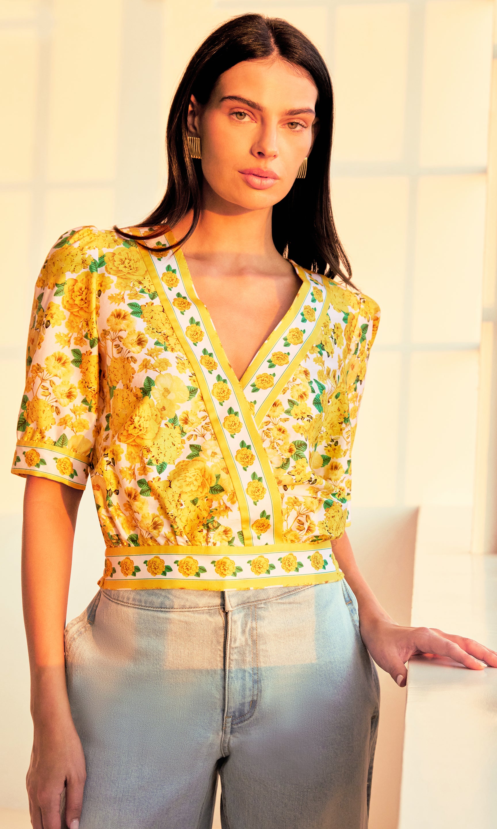 image of model in a sunset-lit room wearing the Rema Floral Poplin Blouse in a yellow floral pattern paired with light blue jeans.