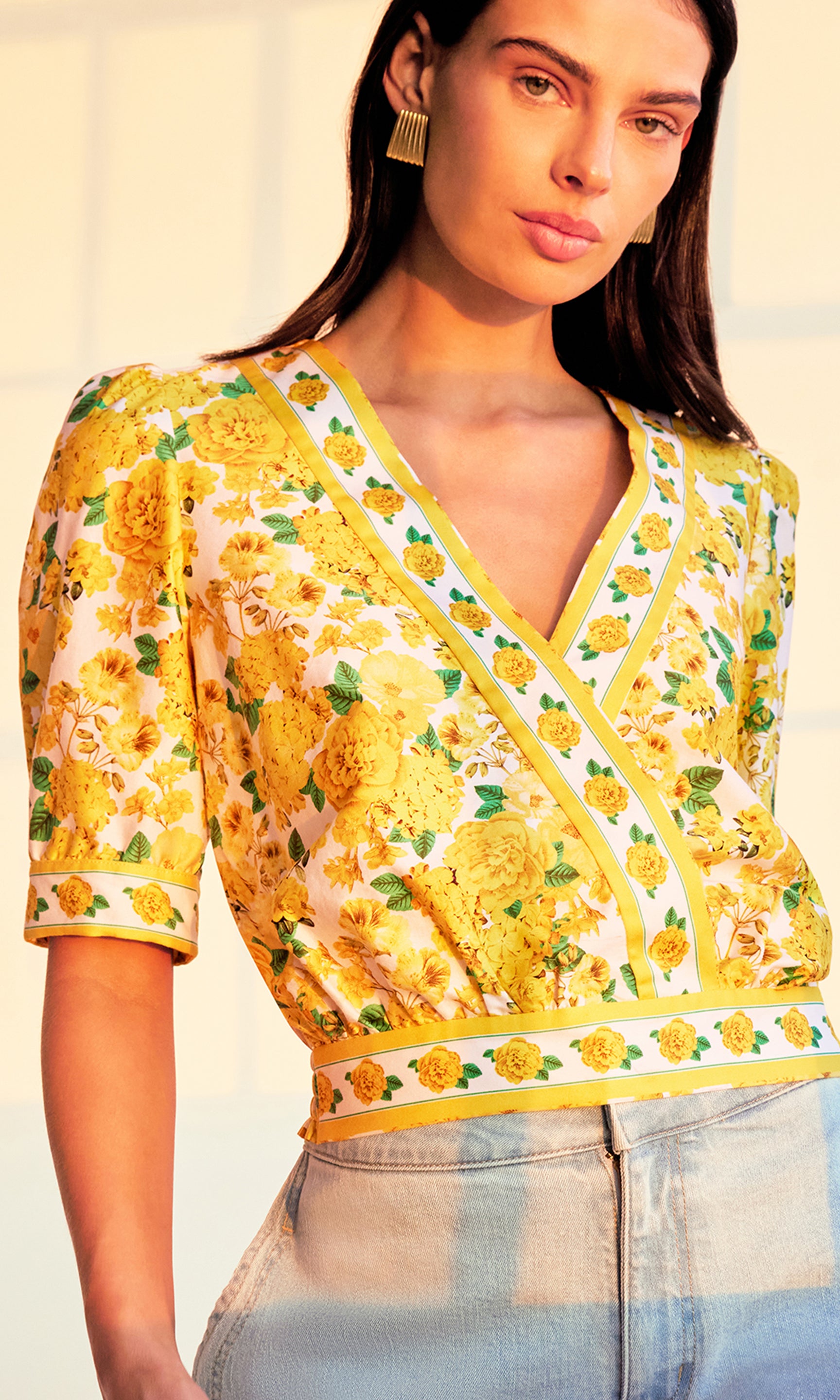 zoomed in image of model in a sunset-lit room wearing the Rema Floral Poplin Blouse in a yellow floral pattern paired showcasing the beautiful tailoring on the blouse with light blue jeans.