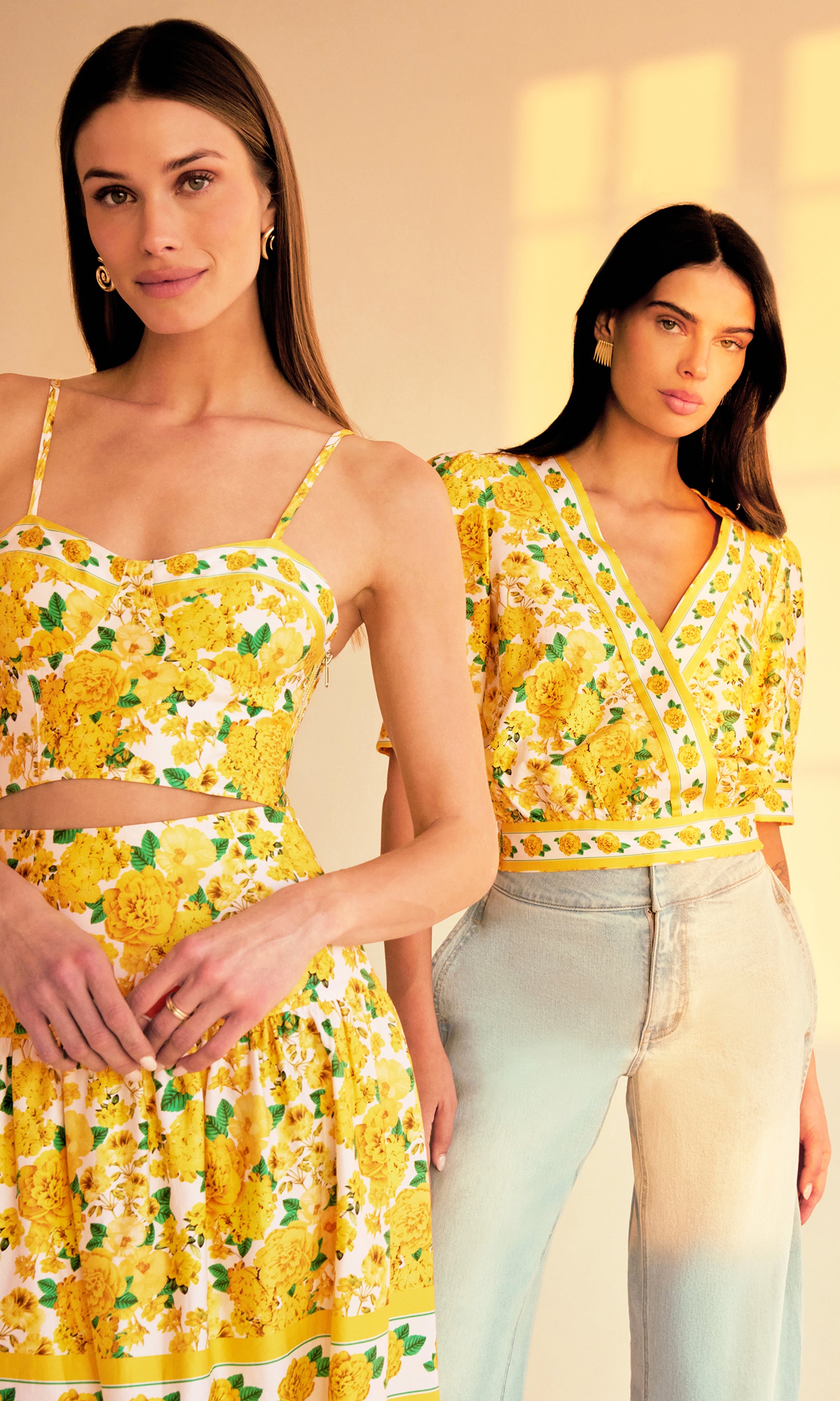 image of two models in a sunset-lit room both wearing styles from the same yellow floral pattern, on the right she is wearing the Rema Blouse with light blue jeans and on the left a bra cami top with a matching maxi skirt.