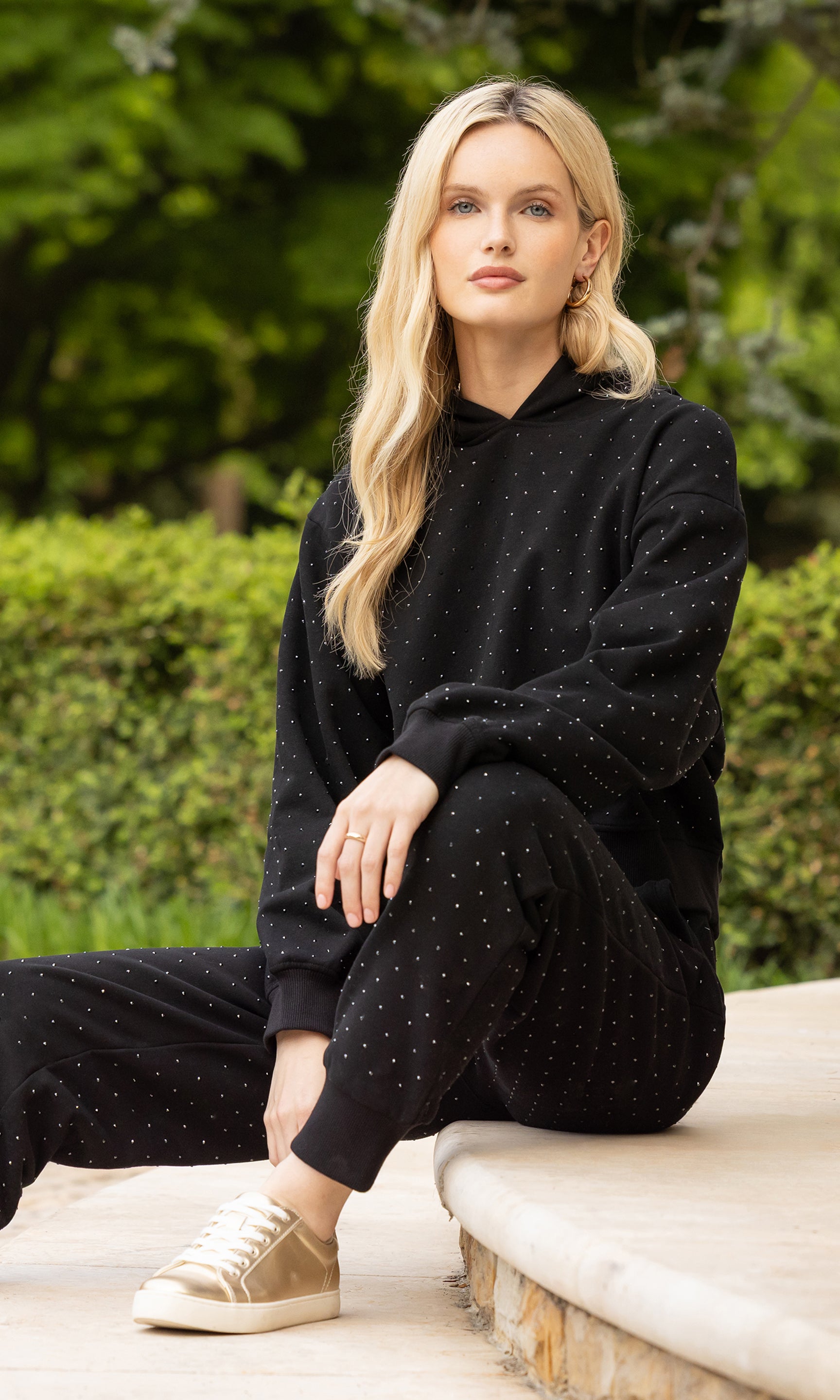 image of model sitting on a step outdoors in a patio wearing a black sweatsuit embellished with crystals and paired with gold sneakers