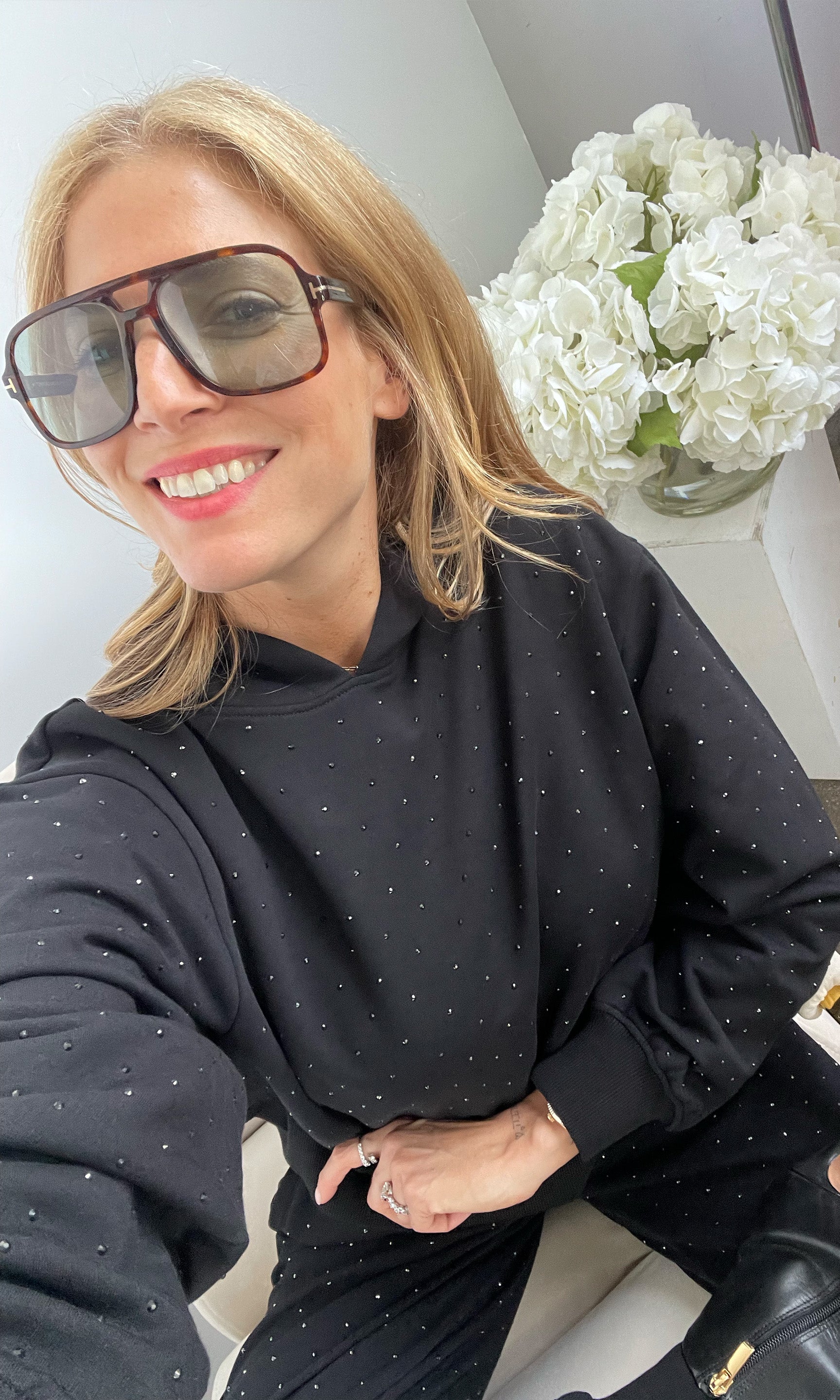 Woman wearing sunglasses and a black polka dot outfit sitting indoors with white flowers in the background.