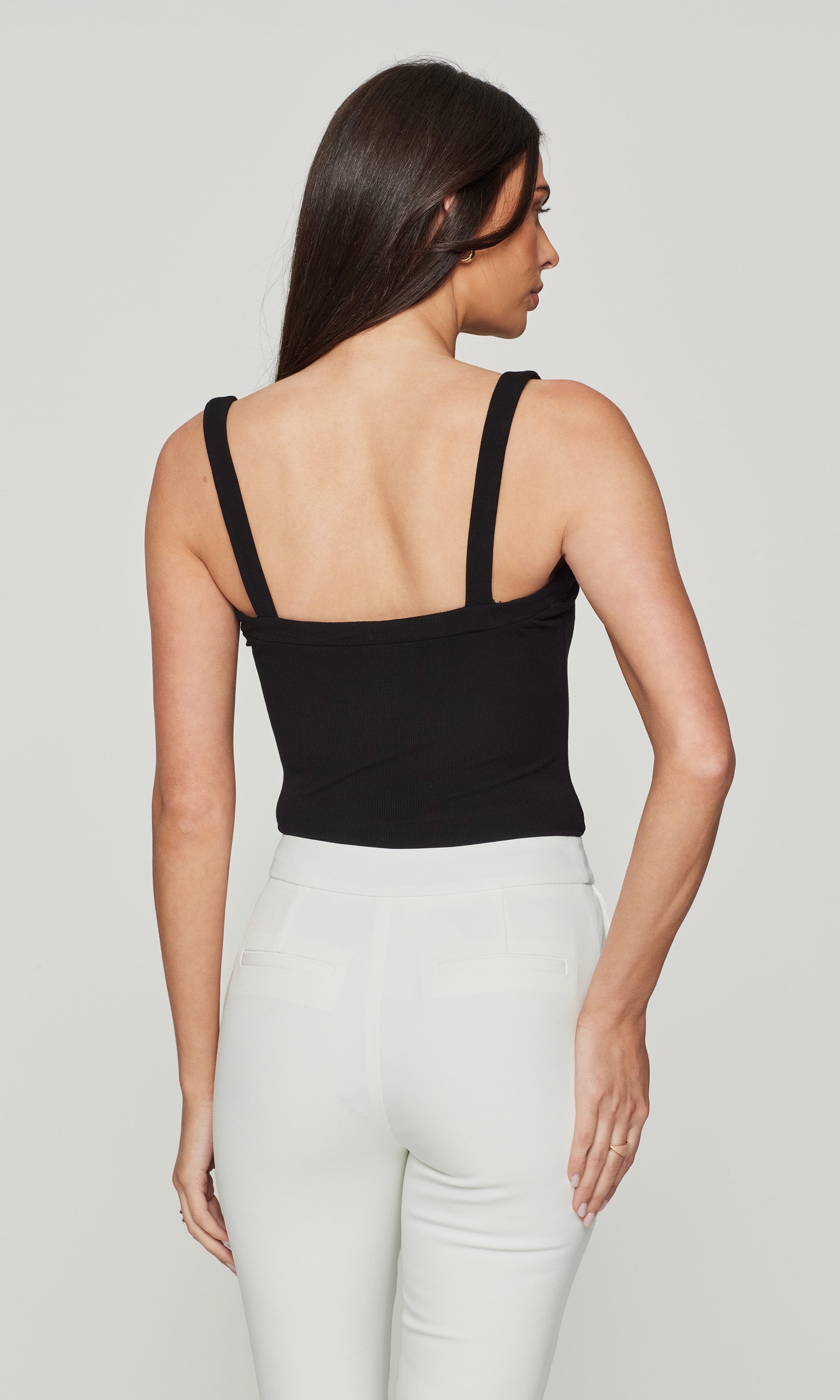 Back view of woman wearing a black tank bodysuit and white pants on a plain background
