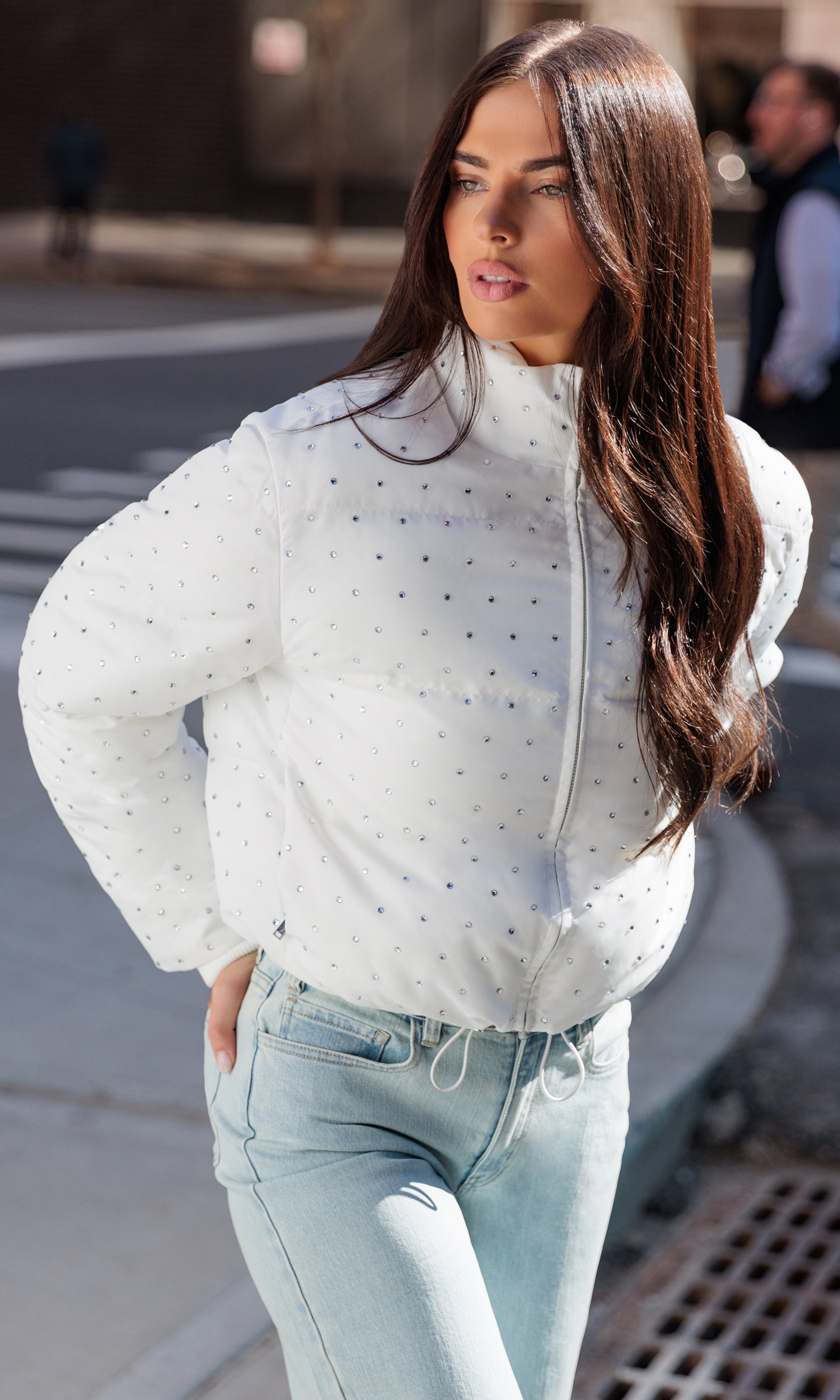 Woman wearing a white crystal embellished jacket and light blue jeans on a city street.