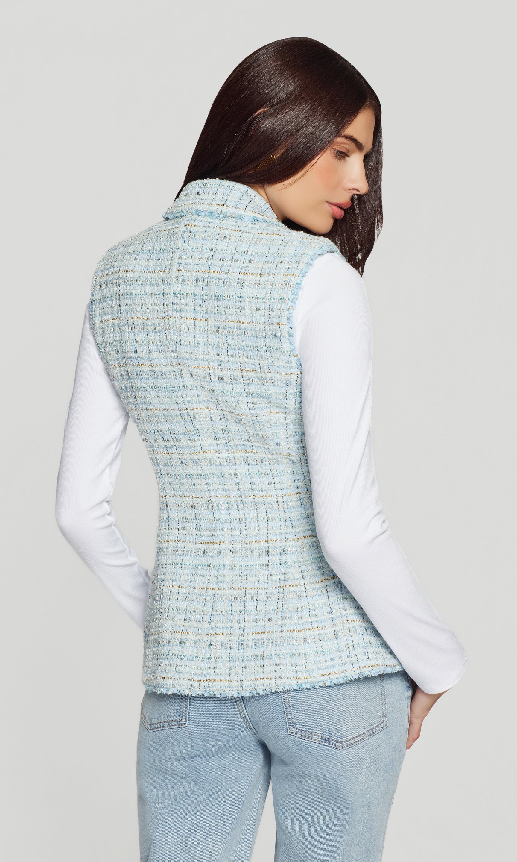 Back view of woman wearing a light blue tweed vest over a white shirt and jeans on a plain background