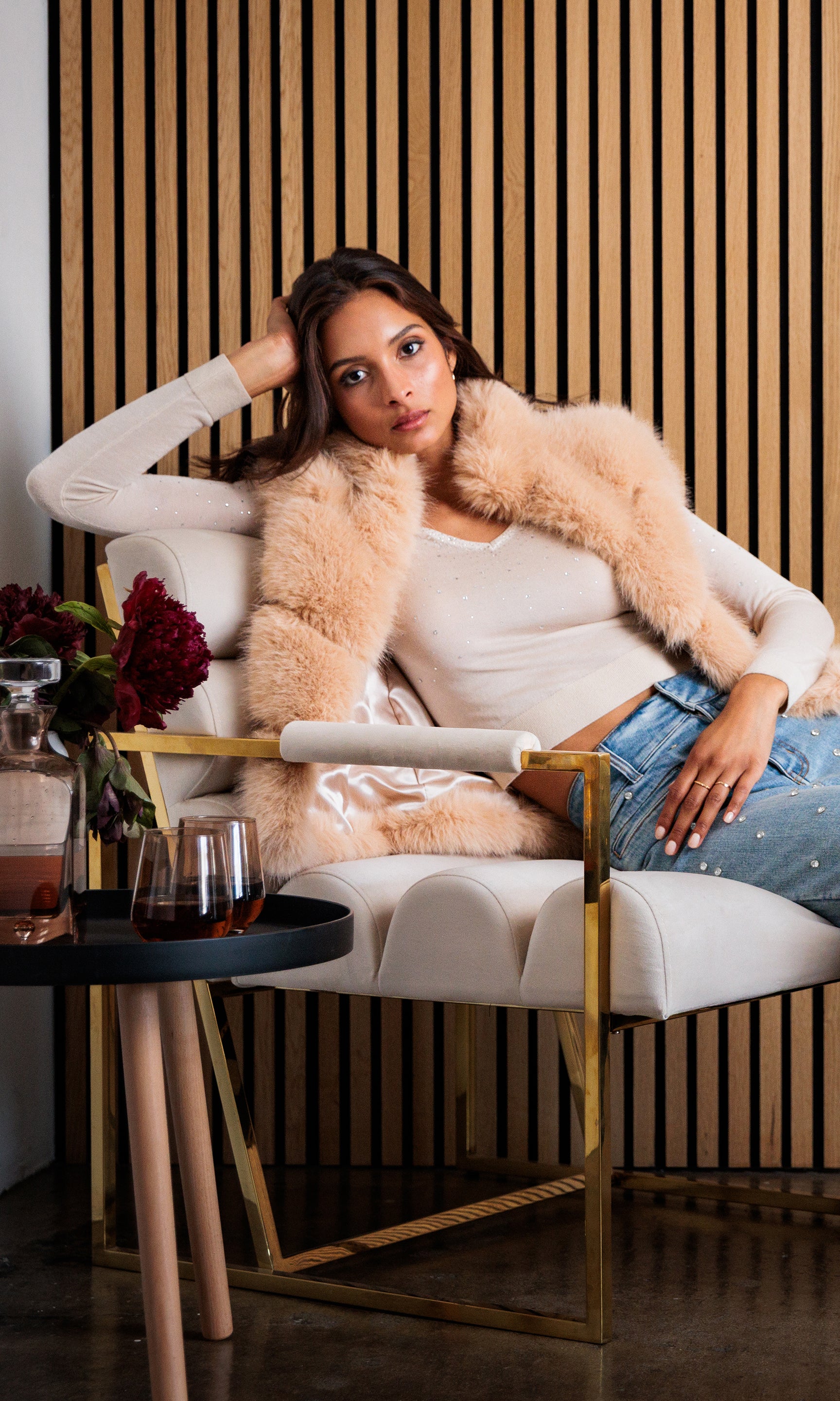 The model sits at a table in a relaxed pose, wearing the Mirasol Crystal Sweater tucked into high-waisted jeans.