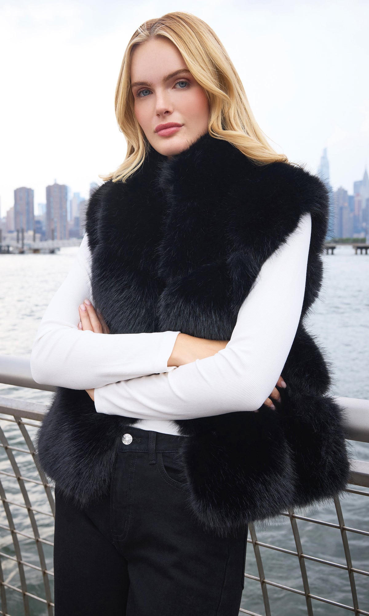 Model wearing a black fur vest over a white long-sleeve top and black pants, standing outdoors with her arms crossed in front of a waterfront city skyline.