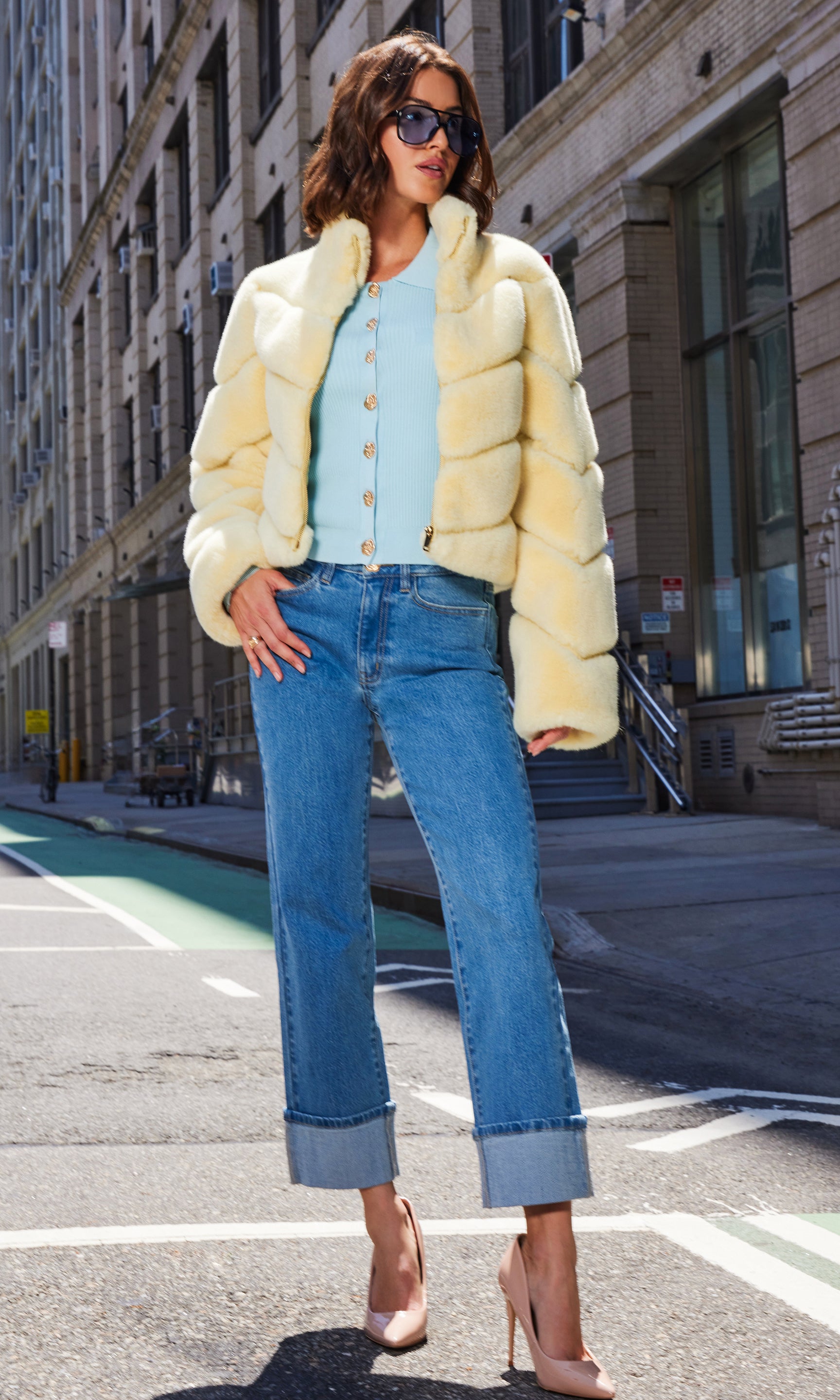 The model strikes a full body view wearing the Brianna Faux Fur Jacket in pale yellow layered over a pastel blue cardigan and slim blue jeans, highlighting its relaxed silhouette as she stands on the sidewalk.